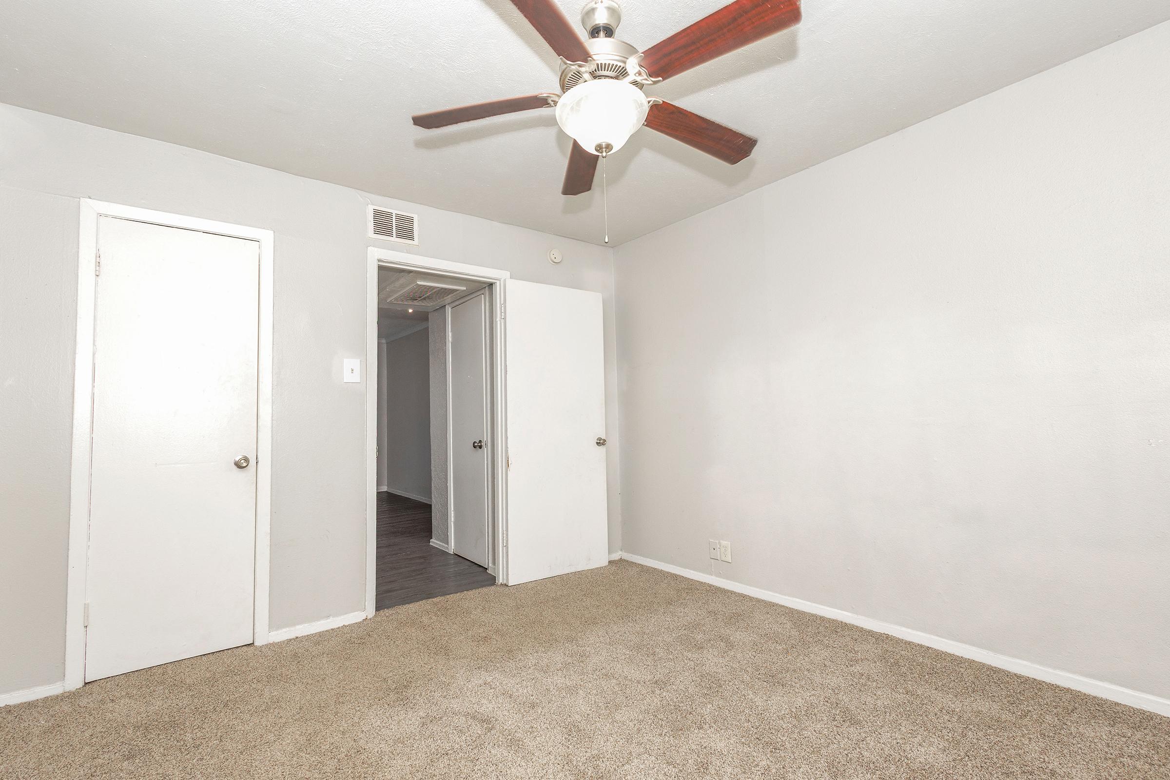 A minimalist room with light gray walls and plush carpet. There's a ceiling fan with wooden blades, and a door on the left leading to another space. Another door is visible in the background, suggesting access to a hallway. The space is well-lit but simple, creating a cozy and spacious atmosphere.