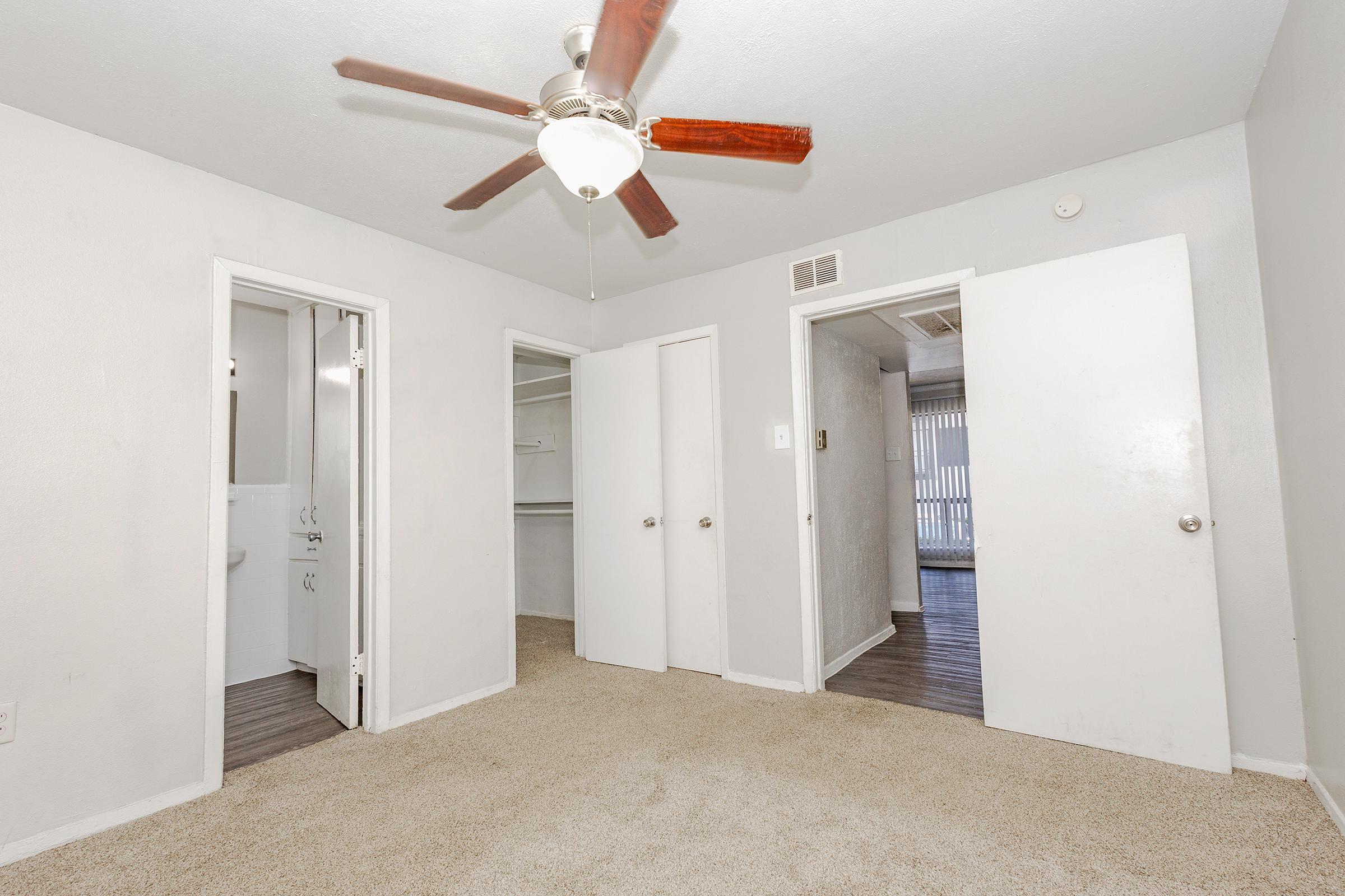 A small, neutral-toned room with light beige carpet and a ceiling fan. Two doors on the left lead to a closet and a bathroom, while another door on the right opens to an outdoor area. The room appears well-lit and spacious, with light-colored walls and a simple, uncluttered layout.