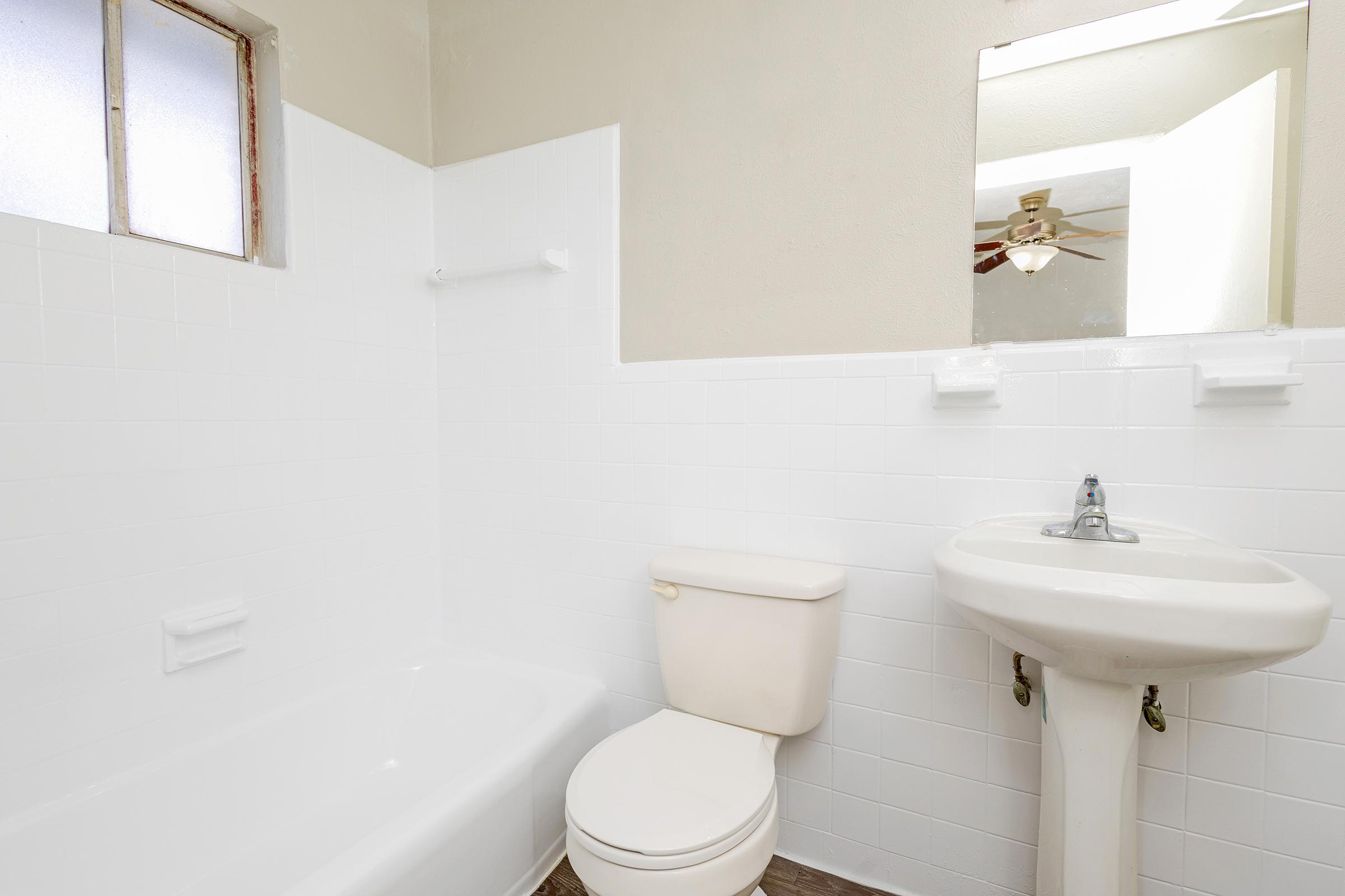 A clean, simple bathroom featuring a white tiled wall, a bathtub with a shower, a toilet, and a small sink. Natural light comes through a window, and a mirror is visible above the sink. The floor appears to be a light wood laminate.