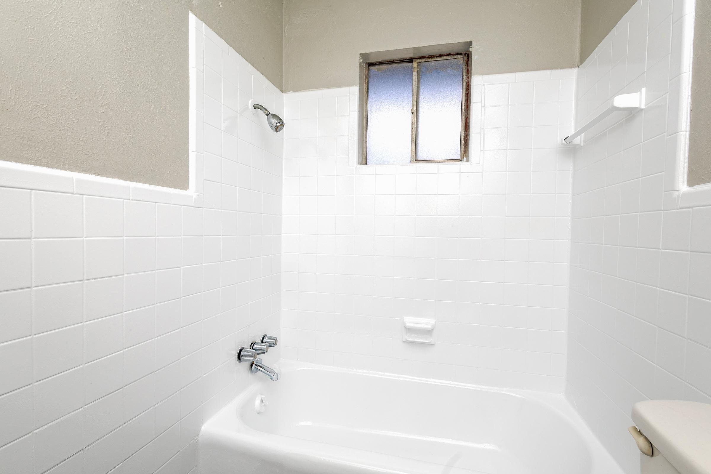 A clean, bright bathroom featuring a white tiled shower and bathtub. There is a showerhead above the tub, a window letting in natural light, and a small shelf for toiletries. The walls are painted in a neutral color, creating a spacious and airy feel.
