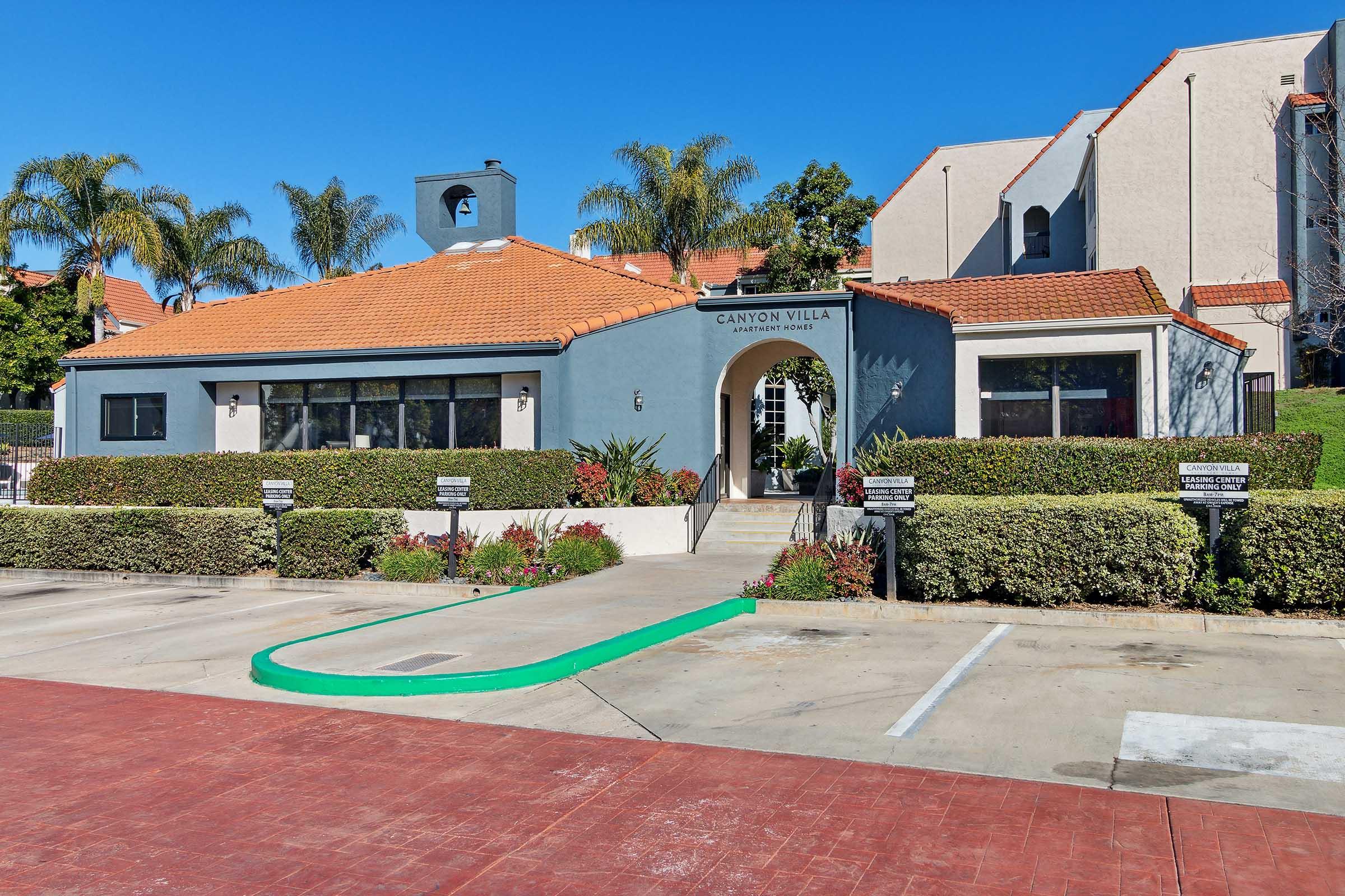 A modern residential building with a pitched orange tile roof, labeled "Canyon Villa." The structure features large windows and a landscaped parking area with green and flowering plants, set against a clear blue sky. Palm trees are visible in the background.