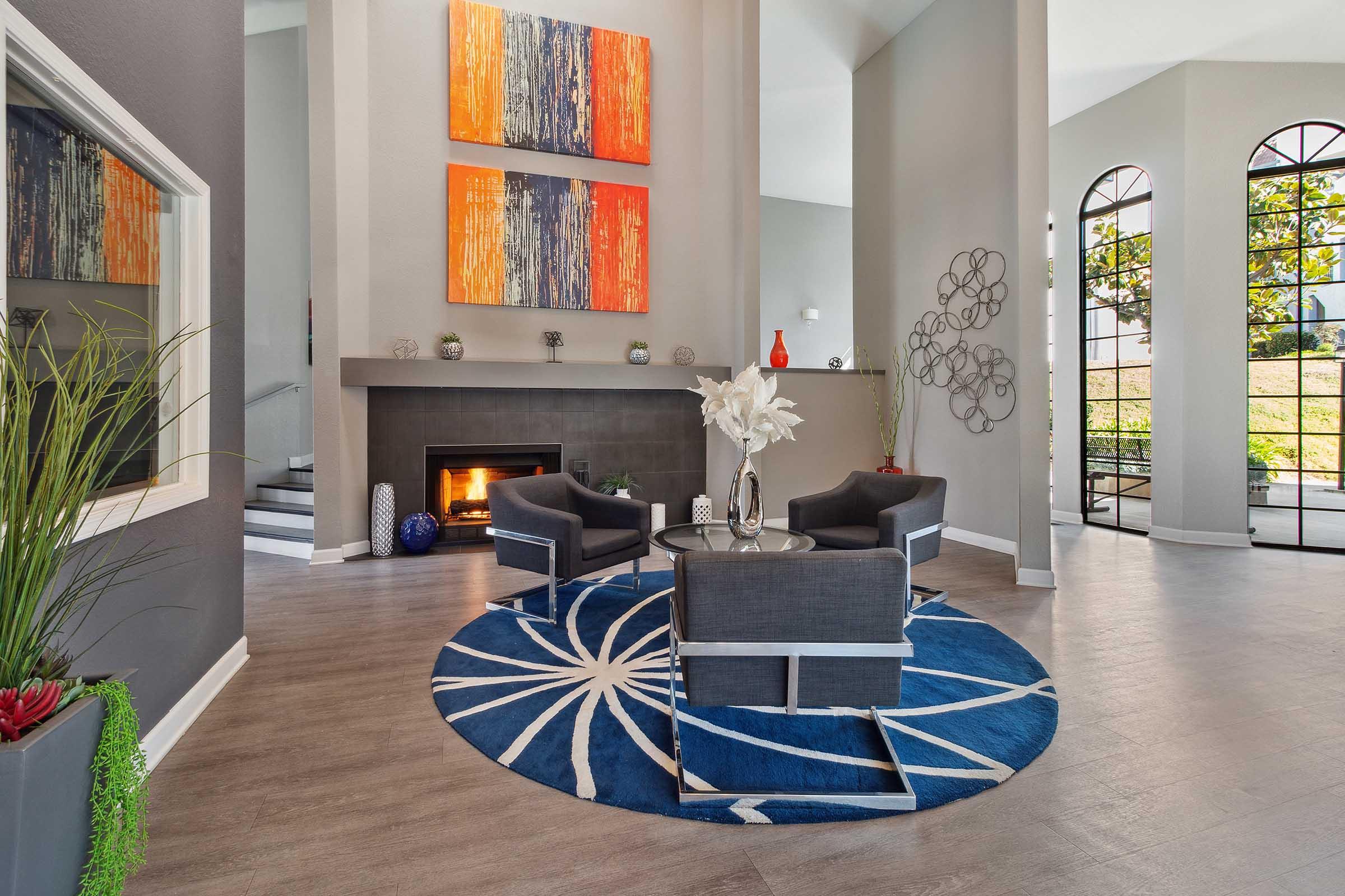 A modern living room featuring a blue circular rug with a white geometric pattern, two black upholstered armchairs around a glass coffee table, and a fireplace. The walls are light gray, adorned with abstract orange and blue artwork. Large windows allow natural light to fill the space, complemented by decorative plants.