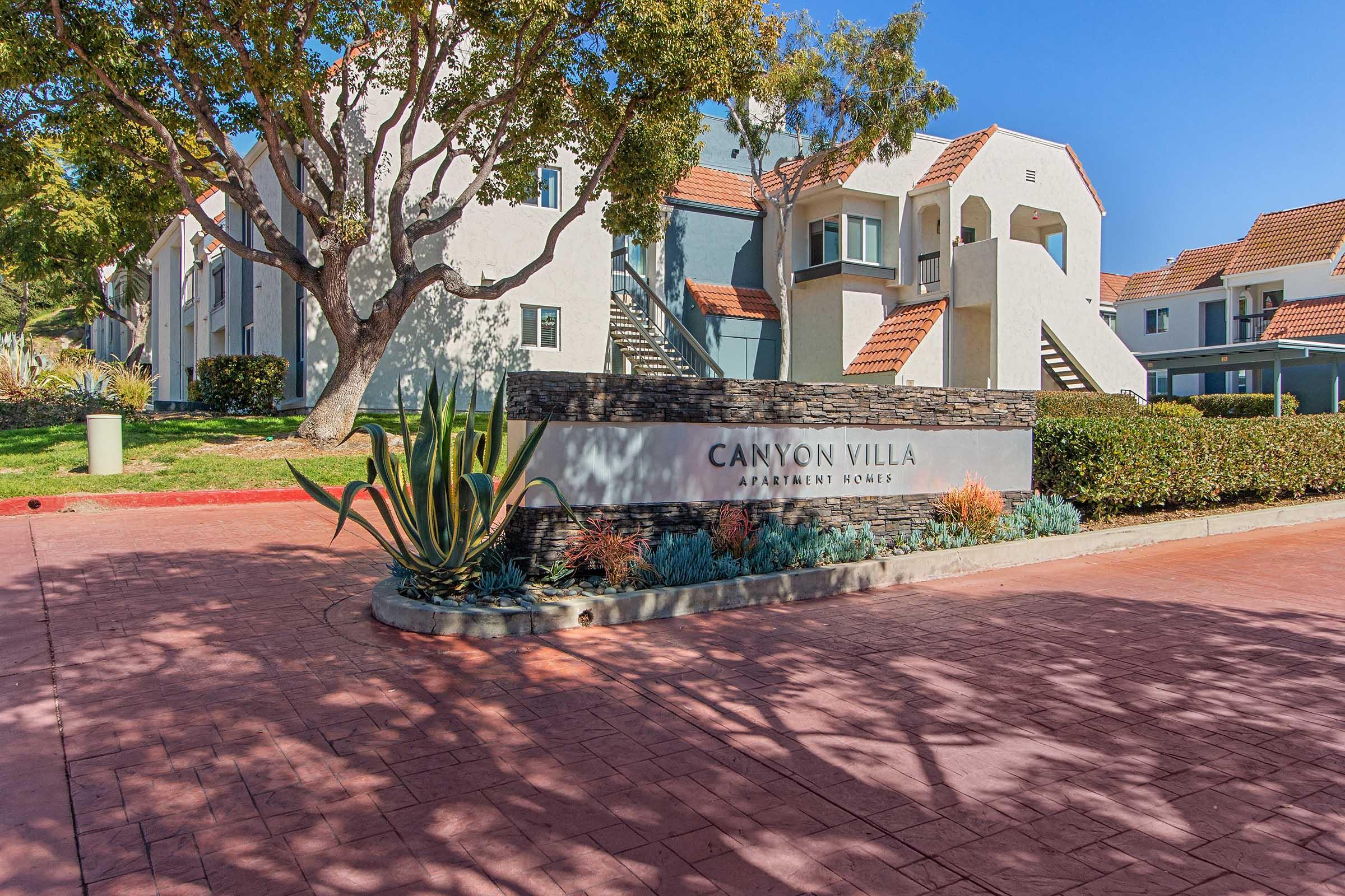 A view of the Canyon Villa Apartment Homes entrance, featuring a stone sign with "Canyon Villa Apartment Homes" displayed prominently. The background includes landscaped greenery, steps leading to apartment entrances, and sunny blue skies. The pathway is paved with red stone, adding to the welcoming atmosphere.