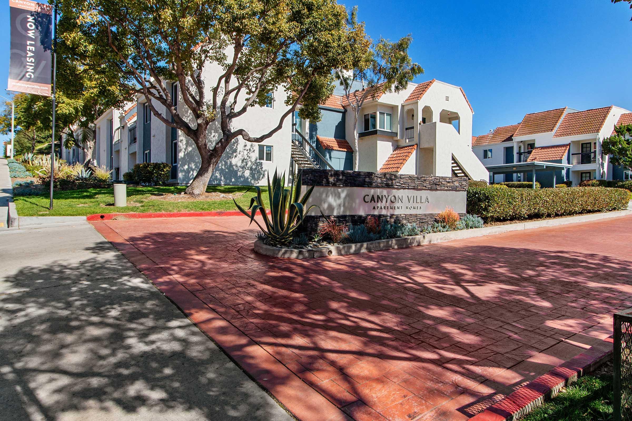 Entrance to Canyon Villa Apartments featuring a well-maintained pathway and greenery. The sign prominently displays "Canyon Villa Apartments," and there are residential buildings visible in the background under a clear blue sky.