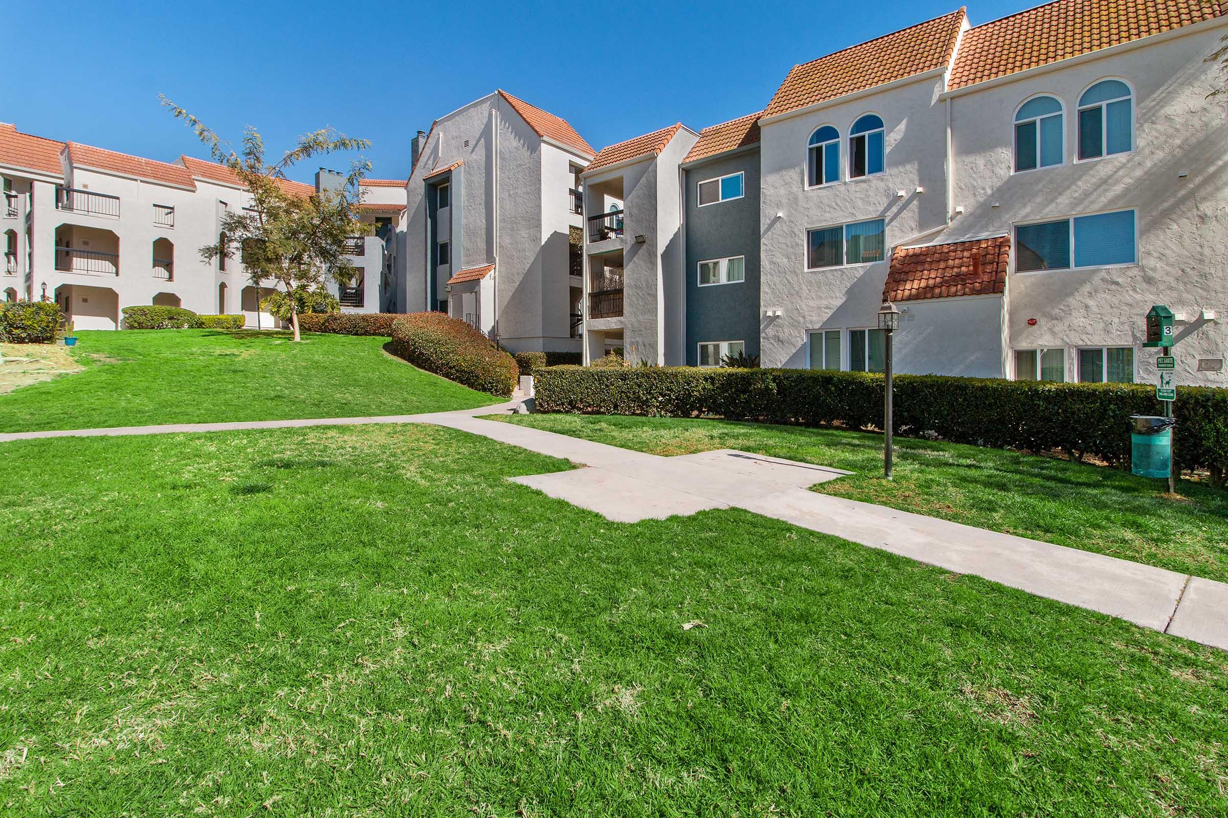 A bright, sunny residential area featuring well-maintained grass and hedges. The scene includes two multistory apartment buildings with red-tiled roofs, surrounded by greenery. A pathway runs through the lawn, leading towards the entrance of the buildings. Clear blue skies enhance the cheerful atmosphere.