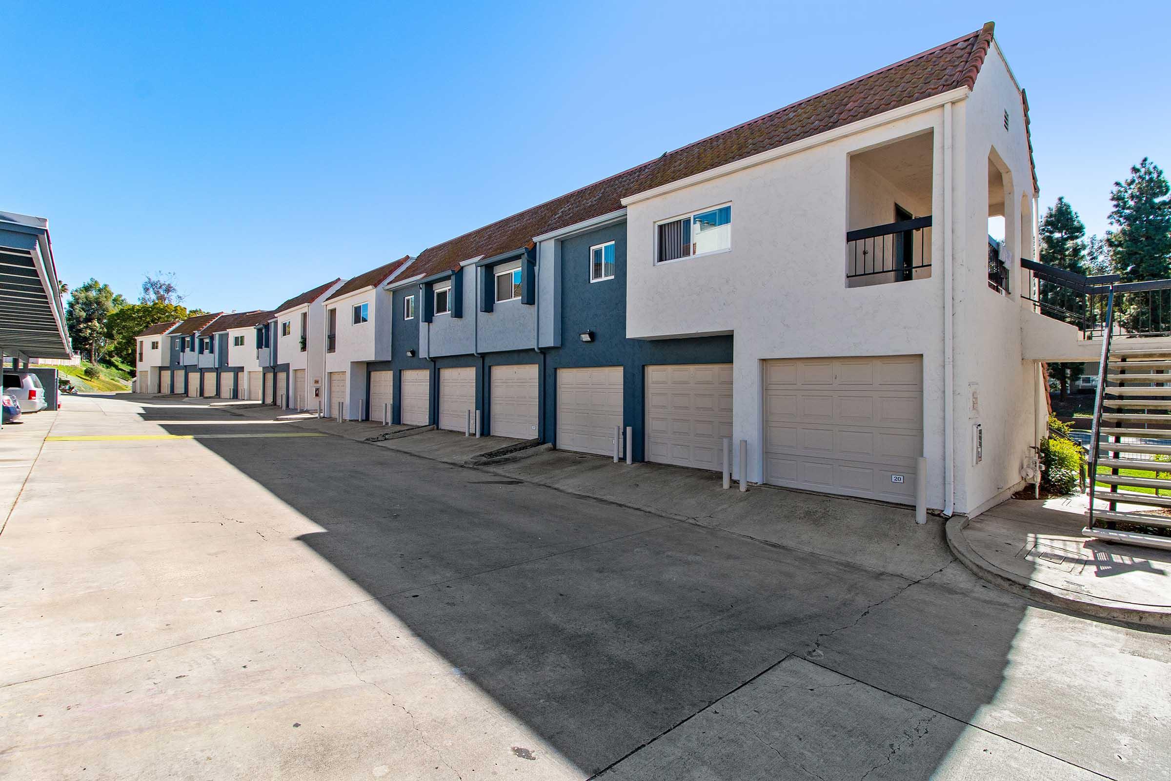 Row of residential buildings with garages at ground level. The architecture features a mix of light colors, including blue and white, and has a clear blue sky overhead. The area is well-maintained with concrete pathways and landscaping alongside the structures.