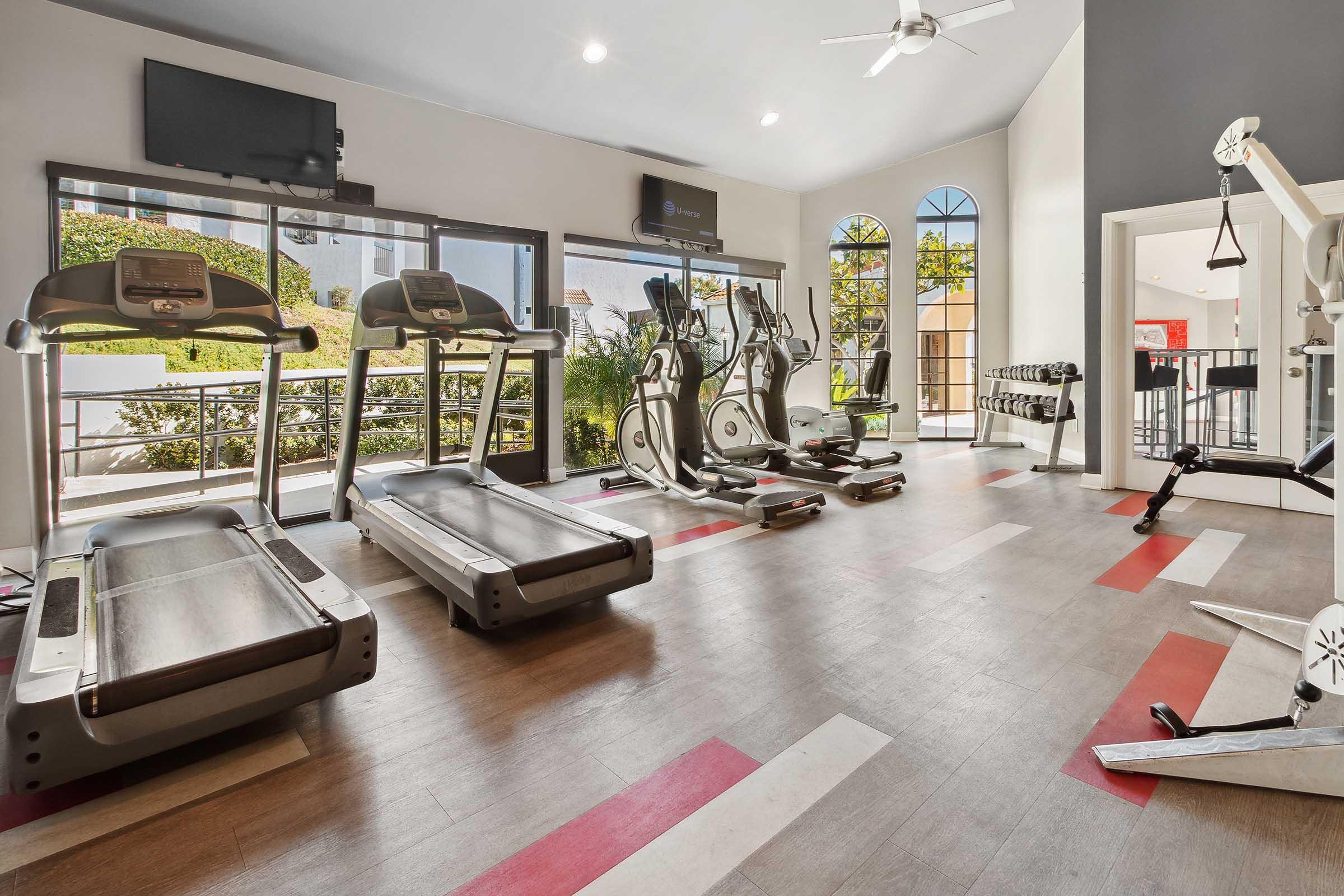 A bright fitness room with multiple treadmills and elliptical machines. Large windows let in natural light, showcasing greenery outside. In the background, there is a small area with resistance machines and a bar-style seating area. The floor has a hardwood appearance with red and white stripes.