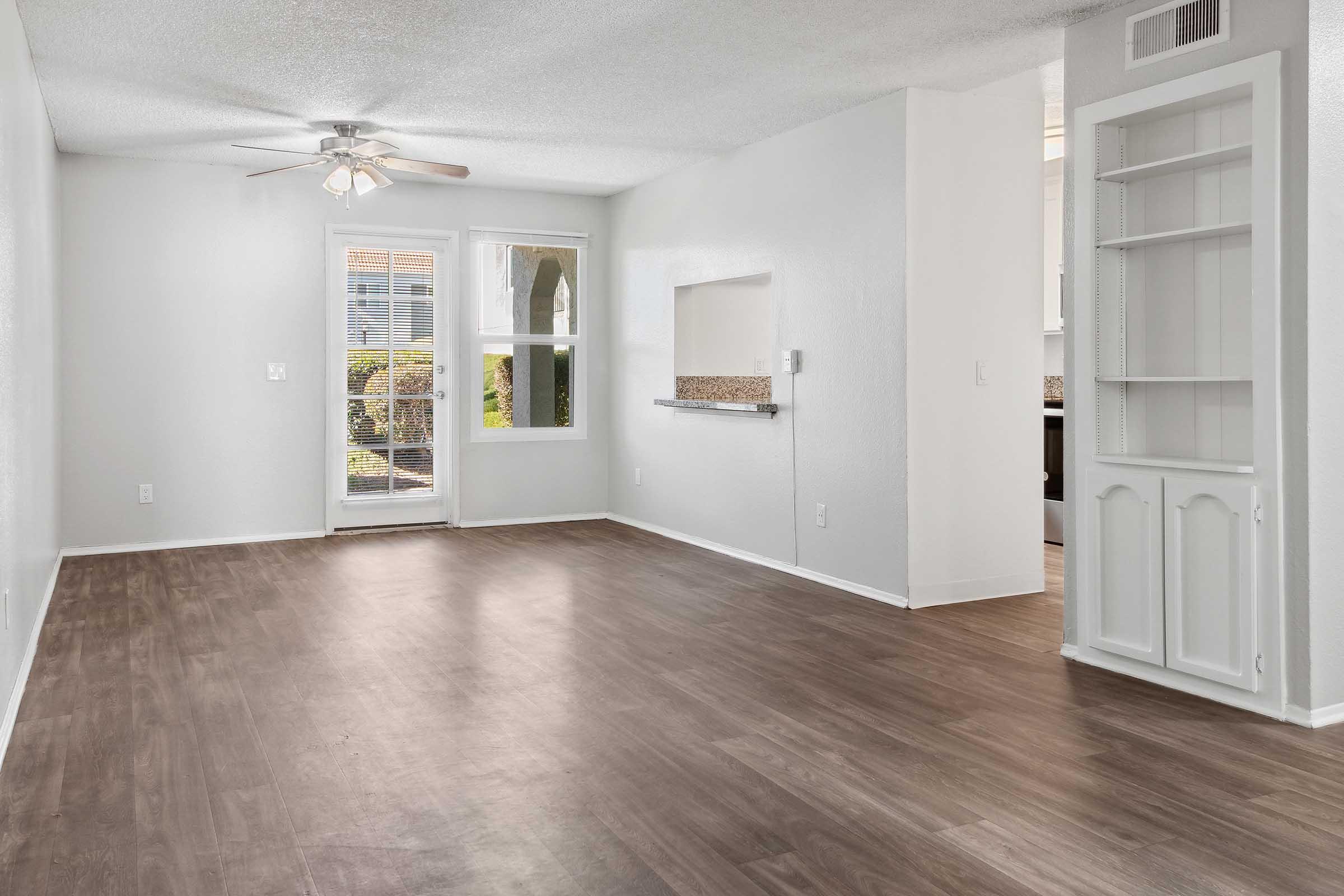 Bright and spacious living room with light-colored walls, large windows, and a ceiling fan. The floor features wood-like laminate. A small alcove with a countertop is visible, along with built-in shelves on one side. The layout promotes an open feel, leading to a kitchen area in the background.