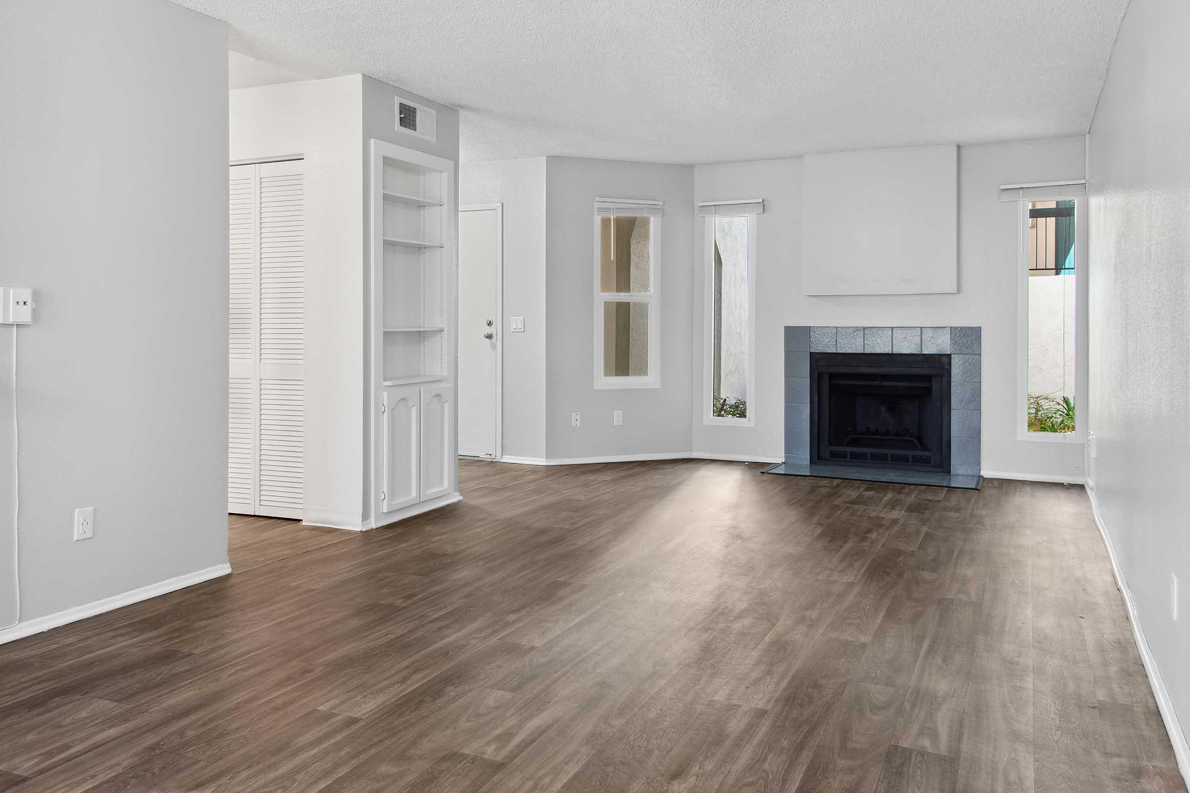Spacious living area featuring light gray walls, laminate flooring, and a decorative fireplace with a slate surround. Natural light enters through large windows, and there is a built-in bookshelf and closet on one wall. The layout is open, providing a welcoming atmosphere.