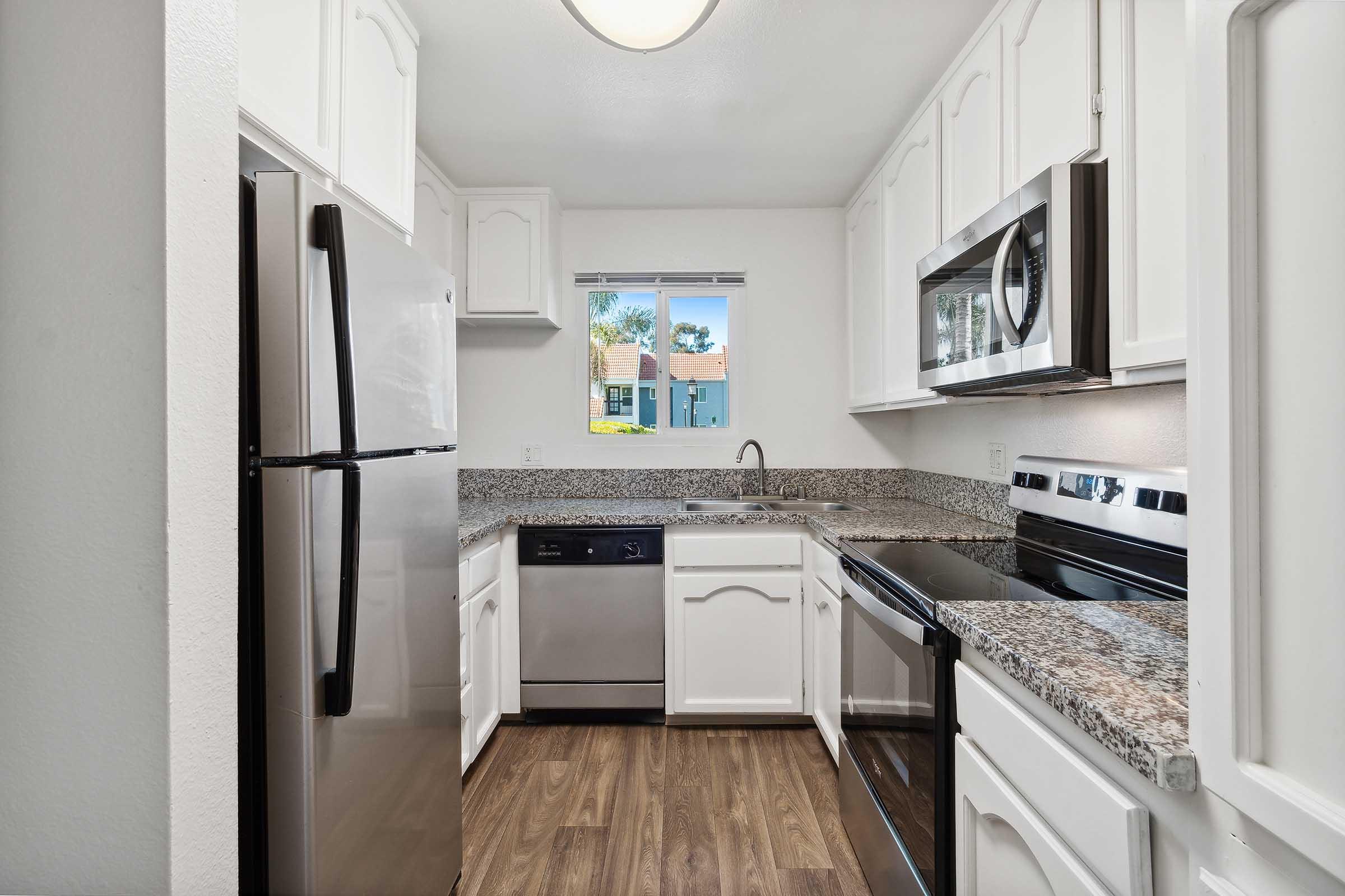 Modern kitchen featuring white cabinets, stainless steel appliances including a refrigerator, dishwasher, and oven, alongside a granite countertop. A window allows natural light, revealing a view of an exterior area. The flooring is a light wood, enhancing the contemporary aesthetic.