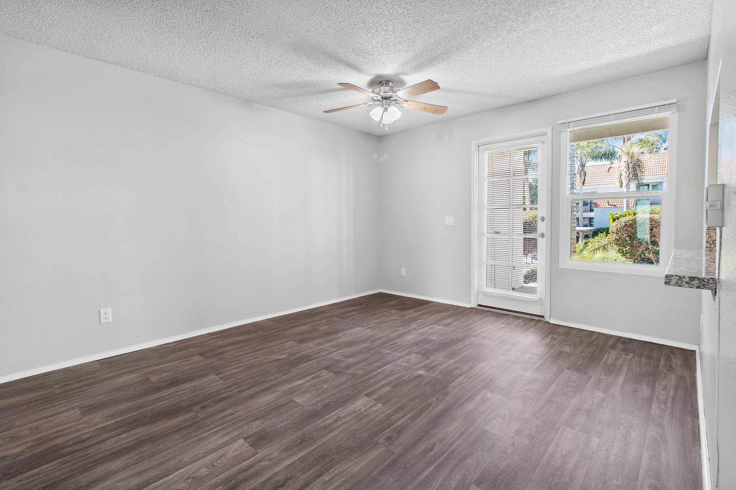 Empty room with light gray walls and a ceiling fan. There is a large window letting in natural light, showcasing palm trees outside. The floor is dark wood laminate, and there is a door leading outside. The space appears clean and ready for furnishing.