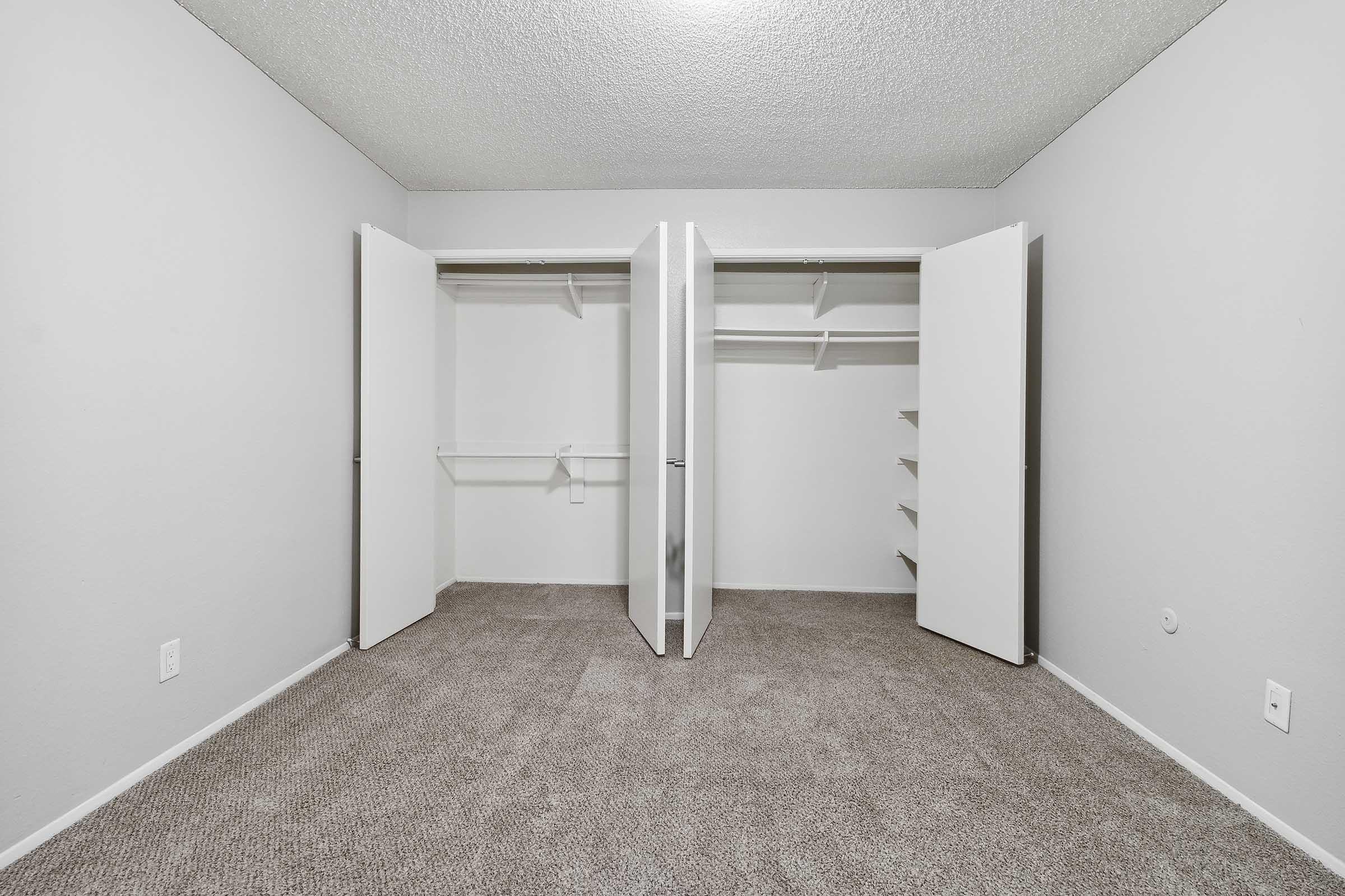 A spacious, empty closet with two bi-fold doors opened, revealing shelving and hanging space inside. The walls and doors are painted white, and the floor is covered with light beige carpet, creating a clean and minimalist appearance.
