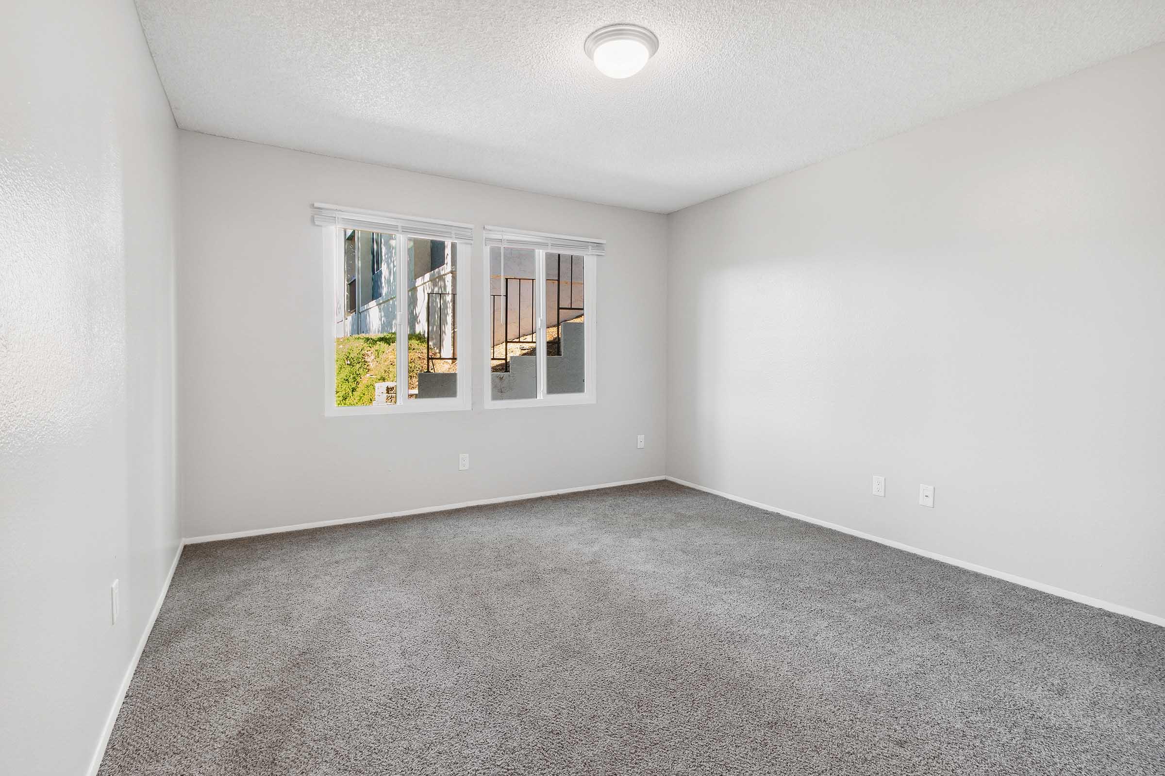 A vacant room with light gray walls and carpet, featuring two windows that allow natural light to enter. The windows have white blinds. There is a ceiling light fixture, and the room appears clean and ready for furnishing.