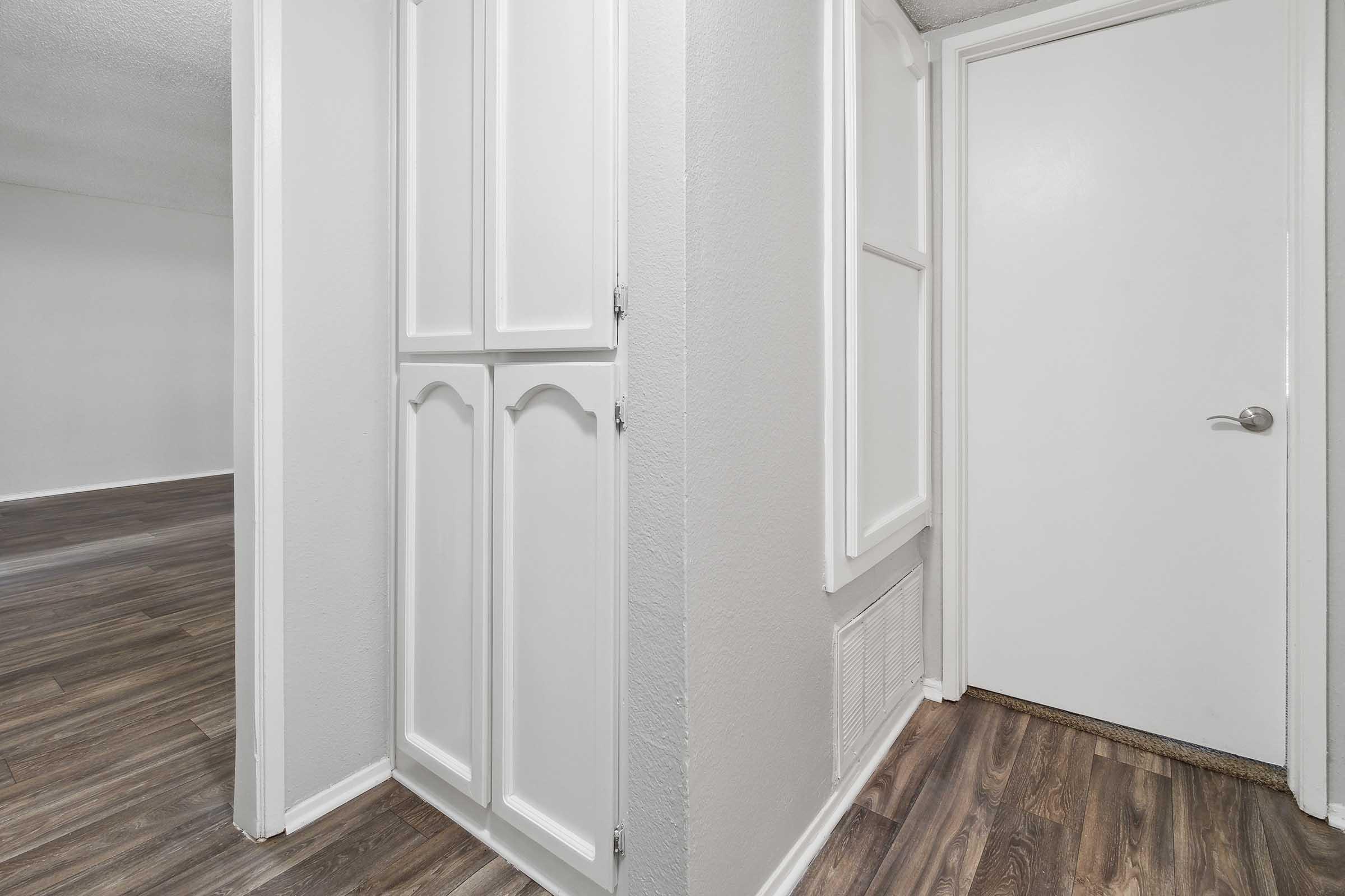A view of a corner in an interior space, featuring a white door on the right and a built-in cabinet with panel doors on the left. The floor is covered with laminate wood, and the walls are painted in light colors, creating a spacious and bright atmosphere.