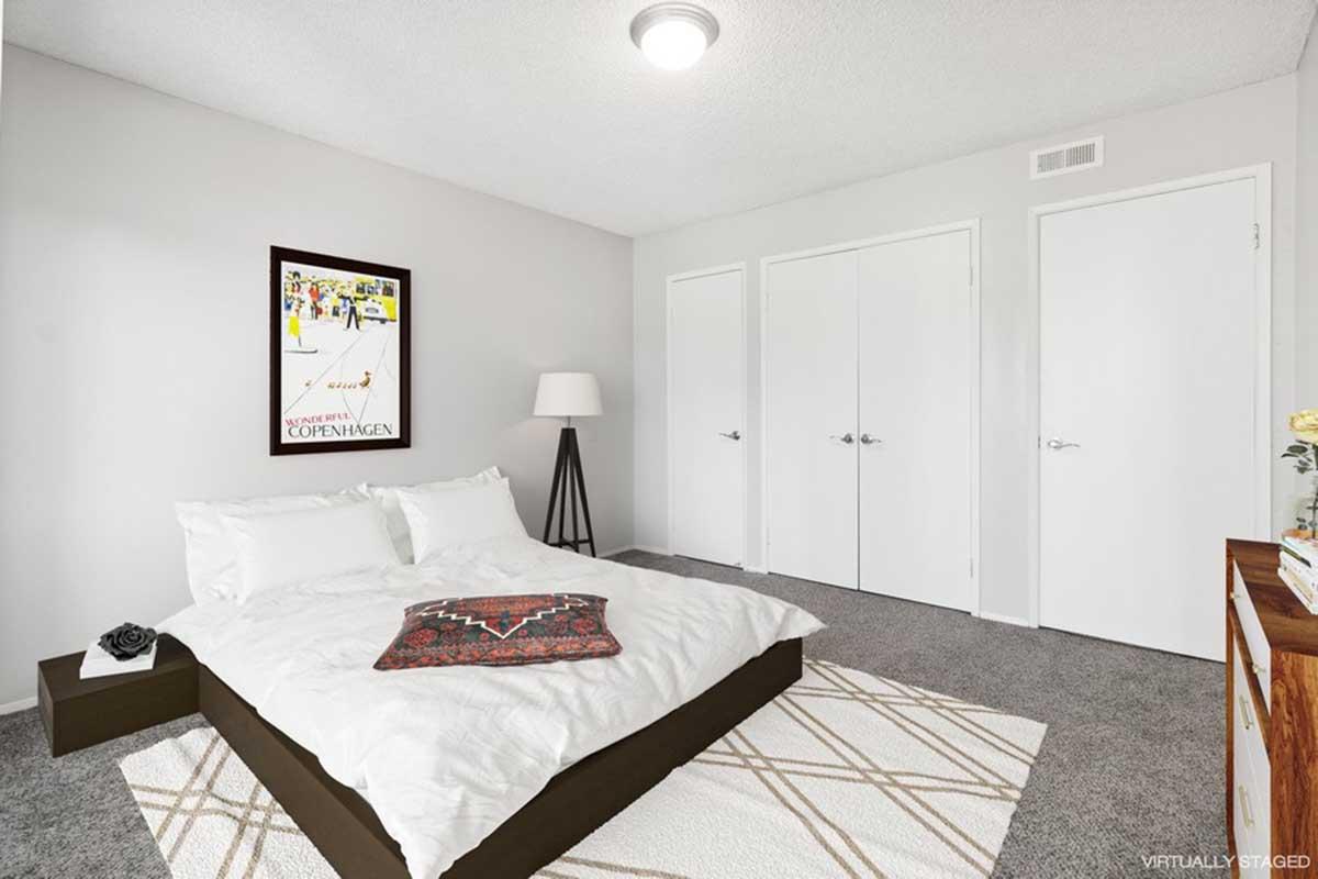 A cozy bedroom featuring a low-profile bed with white bedding and a decorative throw. There's a standing lamp beside the bed, a wall-mounted artwork, and two sliding closet doors. The room has light gray walls and carpet, creating a calm and inviting atmosphere.
