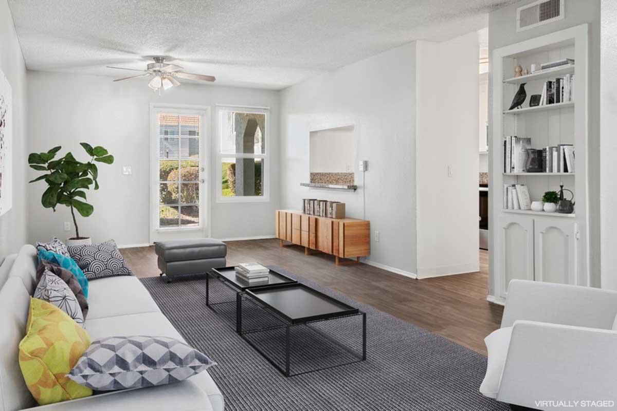 A modern living room featuring a light-colored sofa with colorful cushions, a sleek black coffee table, and a gray area rug. Large windows allow natural light, and there is a decorative plant in the corner. The space has a minimalist design with wooden flooring and a simple entertainment unit.