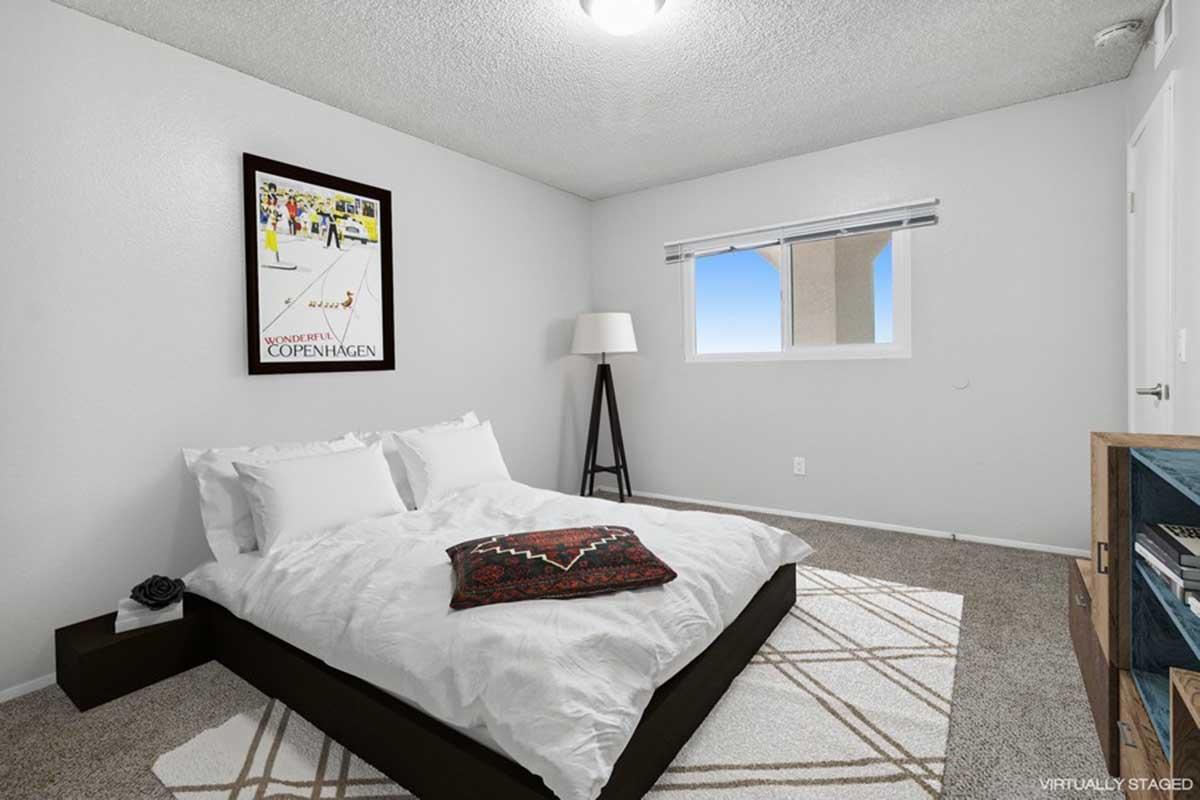 A neatly staged bedroom featuring a bed with white bedding, a decorative throw blanket, and a framed art poster on the wall. There is a floor lamp in the corner and a small wooden side table with an open space for additional decor. A window offers natural light, illuminating the cozy and minimalist design.