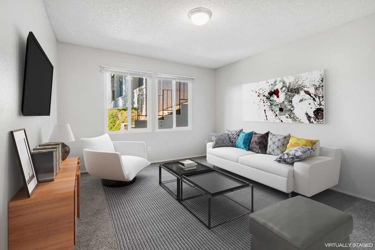A bright, modern living room featuring a white sofa with colorful pillows, a glass coffee table, and a wooden side table. There are two windows allowing natural light, and a large abstract artwork on the wall, with neutral-colored walls and carpet creating a cozy atmosphere.
