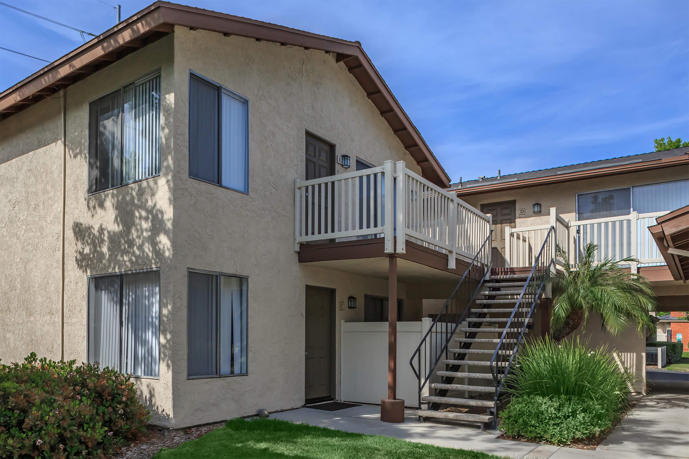 Two-story apartment building with a beige exterior, featuring a balcony on the second floor. A set of outdoor stairs leads to the upper level, and there are large windows with light-colored blinds. Surrounding greenery includes shrubs and grass, creating a welcoming entrance.