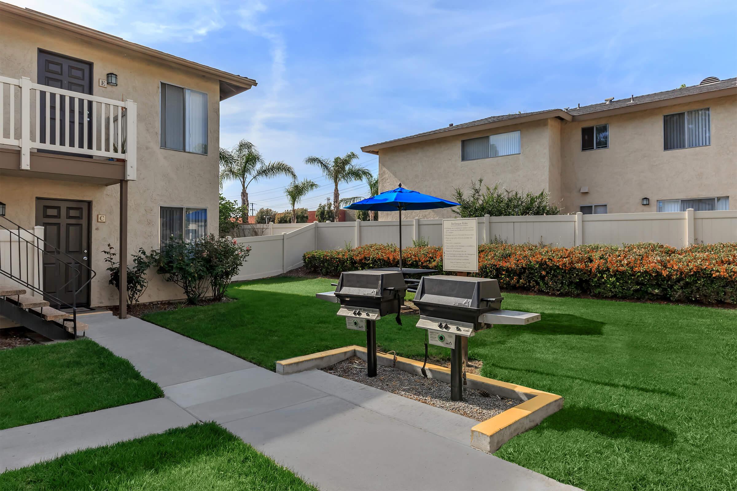 Two outdoor grilling stations are positioned on a paved area between two apartment buildings. The grassy lawn is well-maintained, and there are flower beds with shrubs nearby. A blue umbrella provides shade over a small seating area, creating a welcoming space for residents to relax and enjoy outdoor cooking.