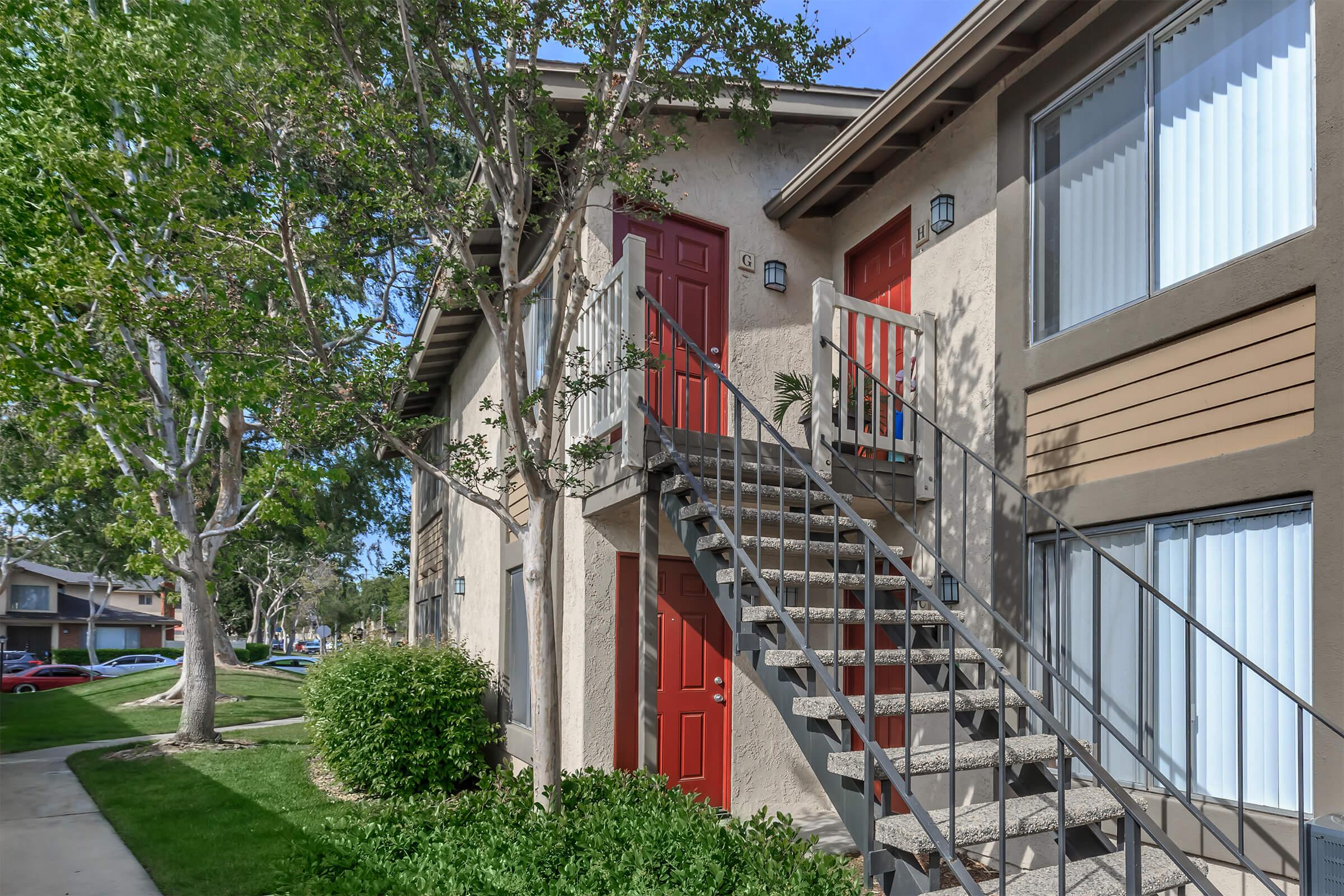 Stairs leading to a second-floor entrance of an apartment building, featuring red doors and decorative light fixtures. Lush green landscaping surrounds the area, with trees providing shade. A walkway leads to the building, and cars are visible parked nearby.