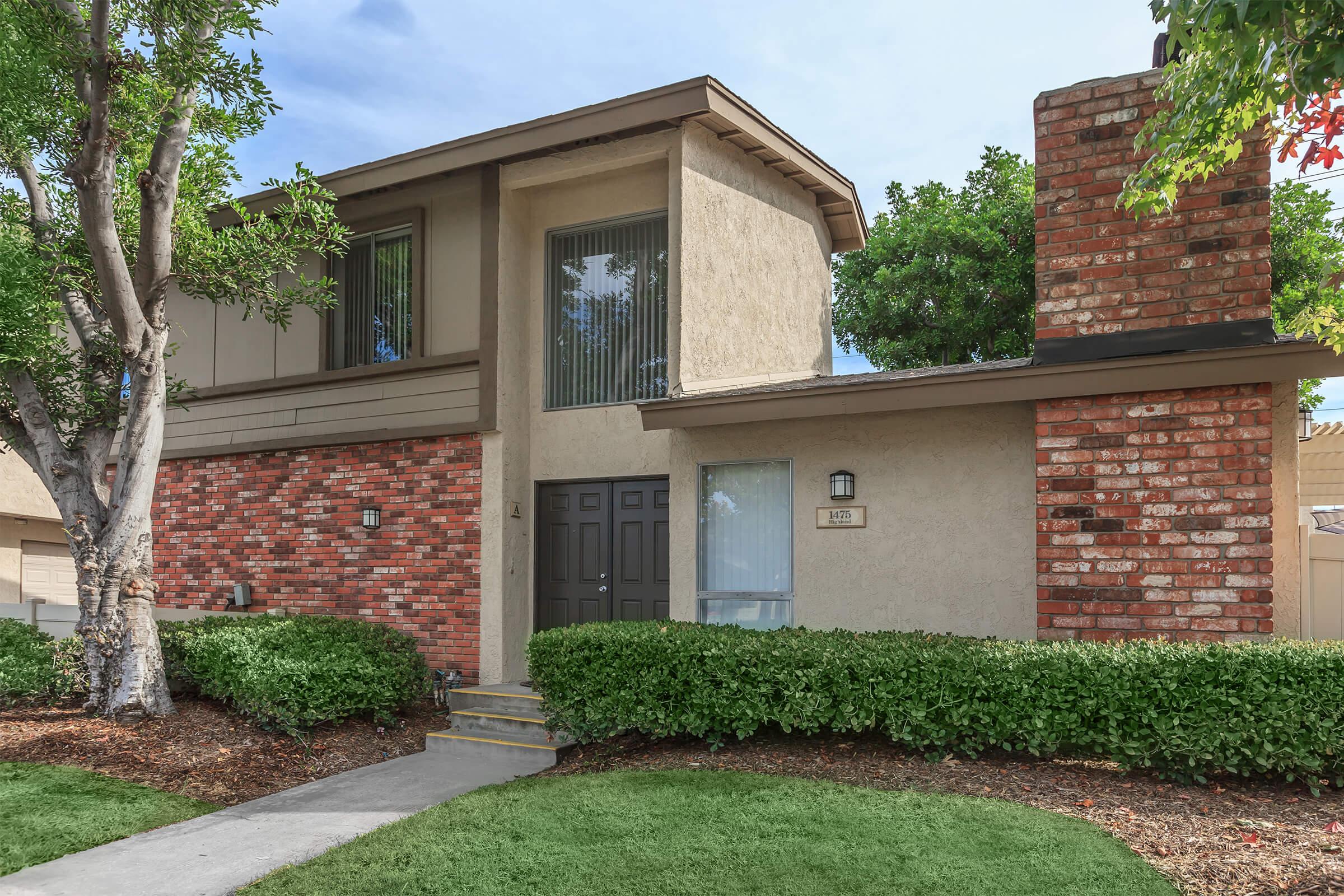 A two-story house featuring a combination of beige stucco and red brick exteriors. It has large windows on the upper level, a front door flanked by decorative wall lighting, and well-maintained shrubbery and lawn. A chimney is visible on the right side, contributing to the home's charm.