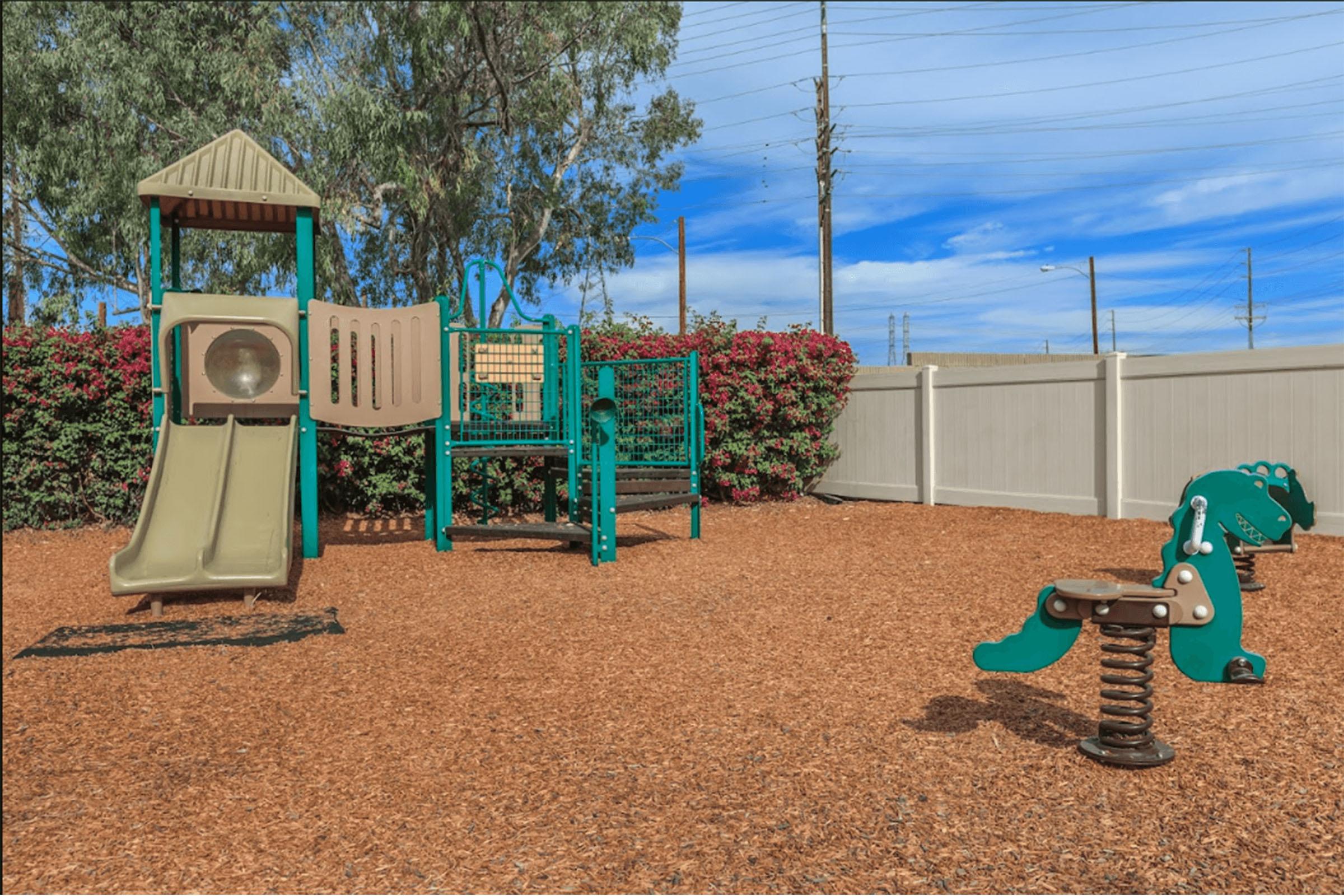 A colorful playground featuring a slide, climbing structure, and a spring rider in the shape of a dinosaur. The area is surrounded by bushes and a white fence, with blue skies and power poles in the background. The ground is covered with soft wood mulch.