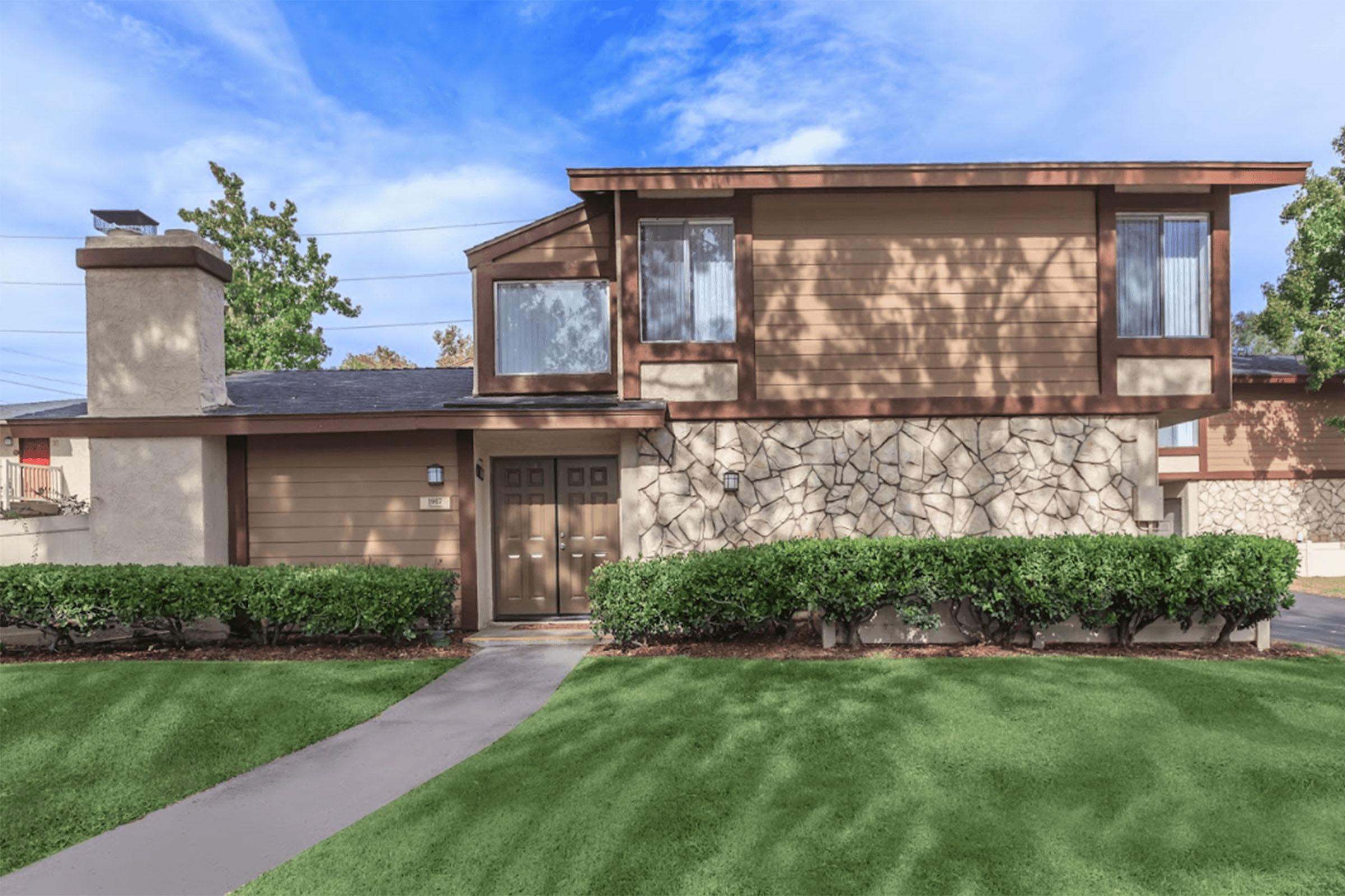 A two-story house with a brown and beige exterior, featuring a stone accent wall and large windows. The front yard has well-trimmed green shrubs and a concrete pathway leading to the entrance. The sky is blue with scattered clouds, and the house is surrounded by a neat lawn.