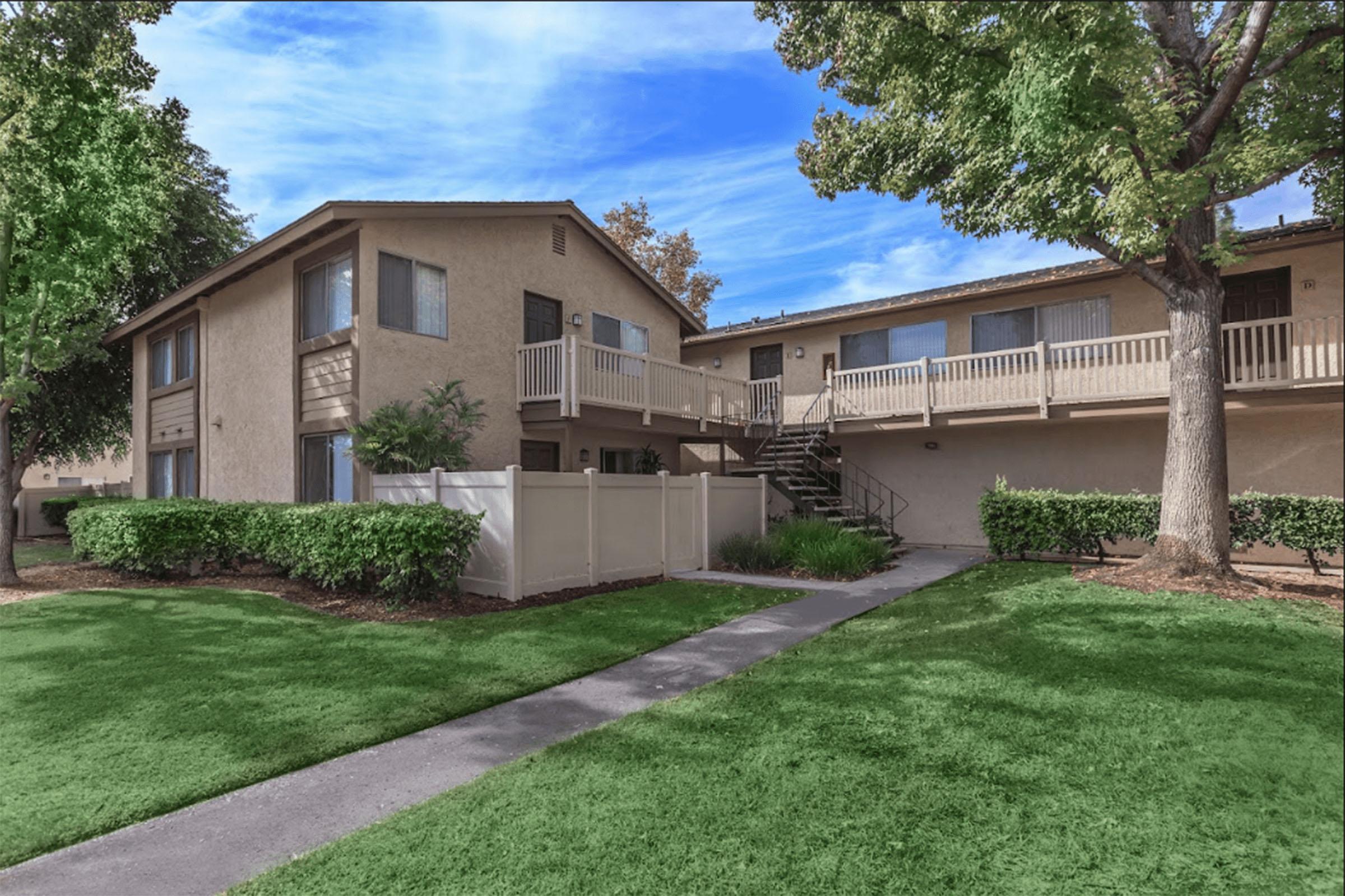 Two-story residential building with beige exterior, featuring balconies on the upper level. The front yard is well-maintained with green grass and shrubs. A paved walkway leads to the entrance, surrounded by trees. Clear blue sky overhead enhances the inviting atmosphere of the property.