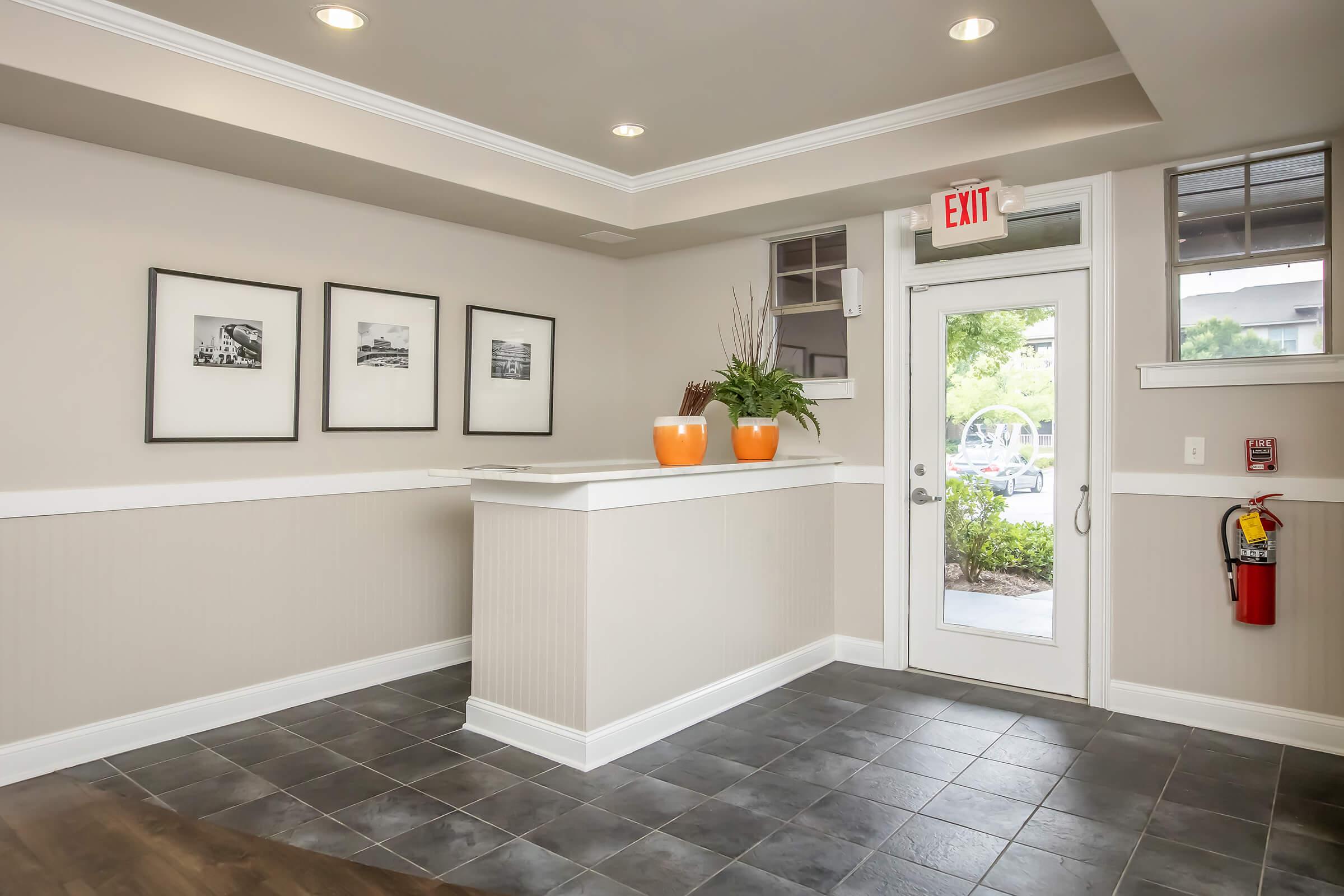 Interior view of a reception area featuring a front desk with two large orange planters, framed black and white photographs on the wall, and a door leading outside. The space is well-lit with recessed lighting, and there is a fire extinguisher mounted on the wall.