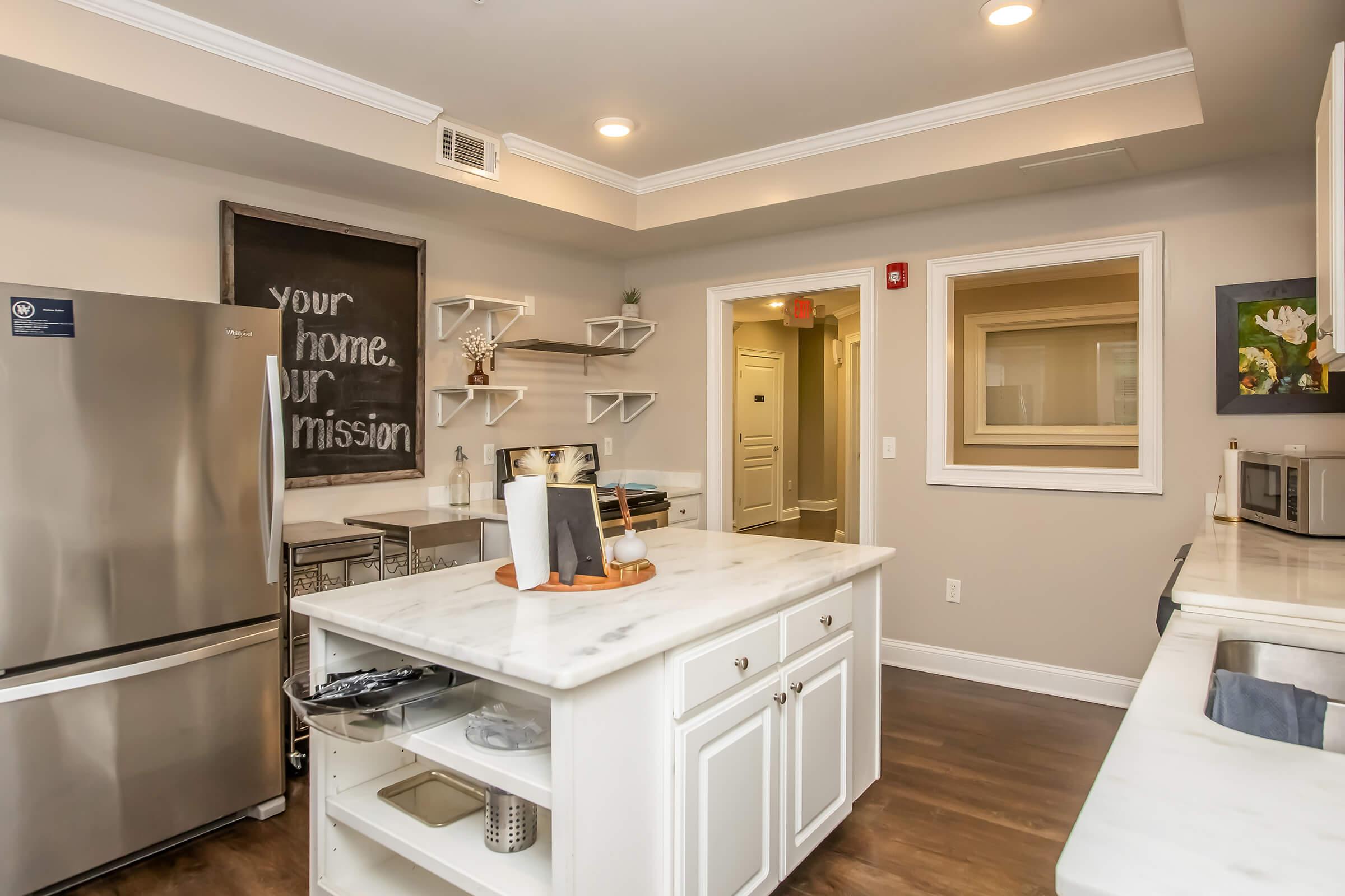 A modern kitchen with white cabinetry and a marble countertop island. Stainless steel fridge and microwave are visible. Open shelving displays decorative items. A chalkboard on the wall reads "your home, our mission." Wood flooring adds warmth, and a door leads to a hallway. Bright lighting illuminates the space.