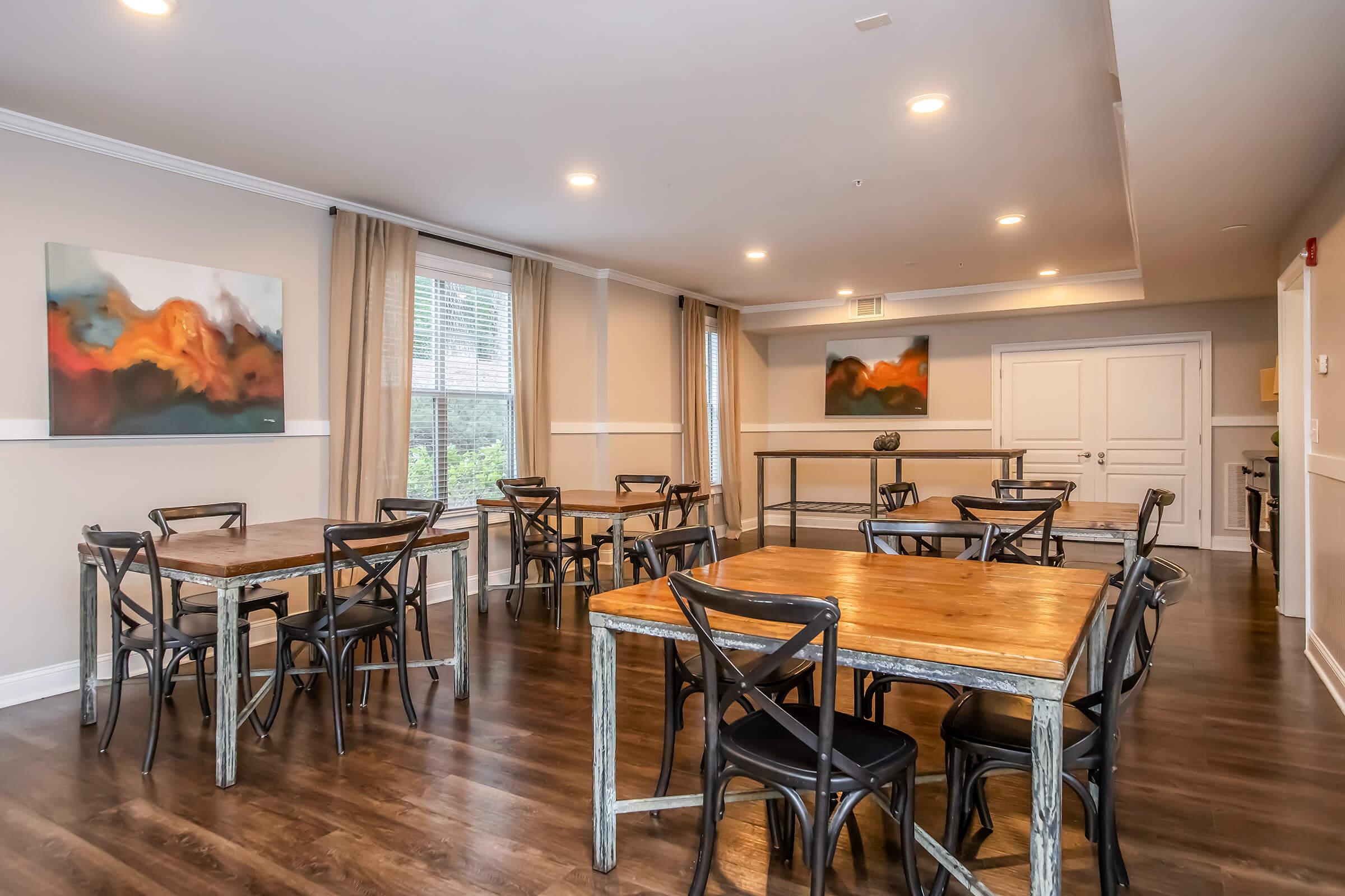 A spacious, well-lit dining room featuring several wooden tables and black chairs. The walls are adorned with abstract paintings. Large windows provide natural light, and there are neutral-colored curtains. The space looks inviting and suitable for gatherings or events.