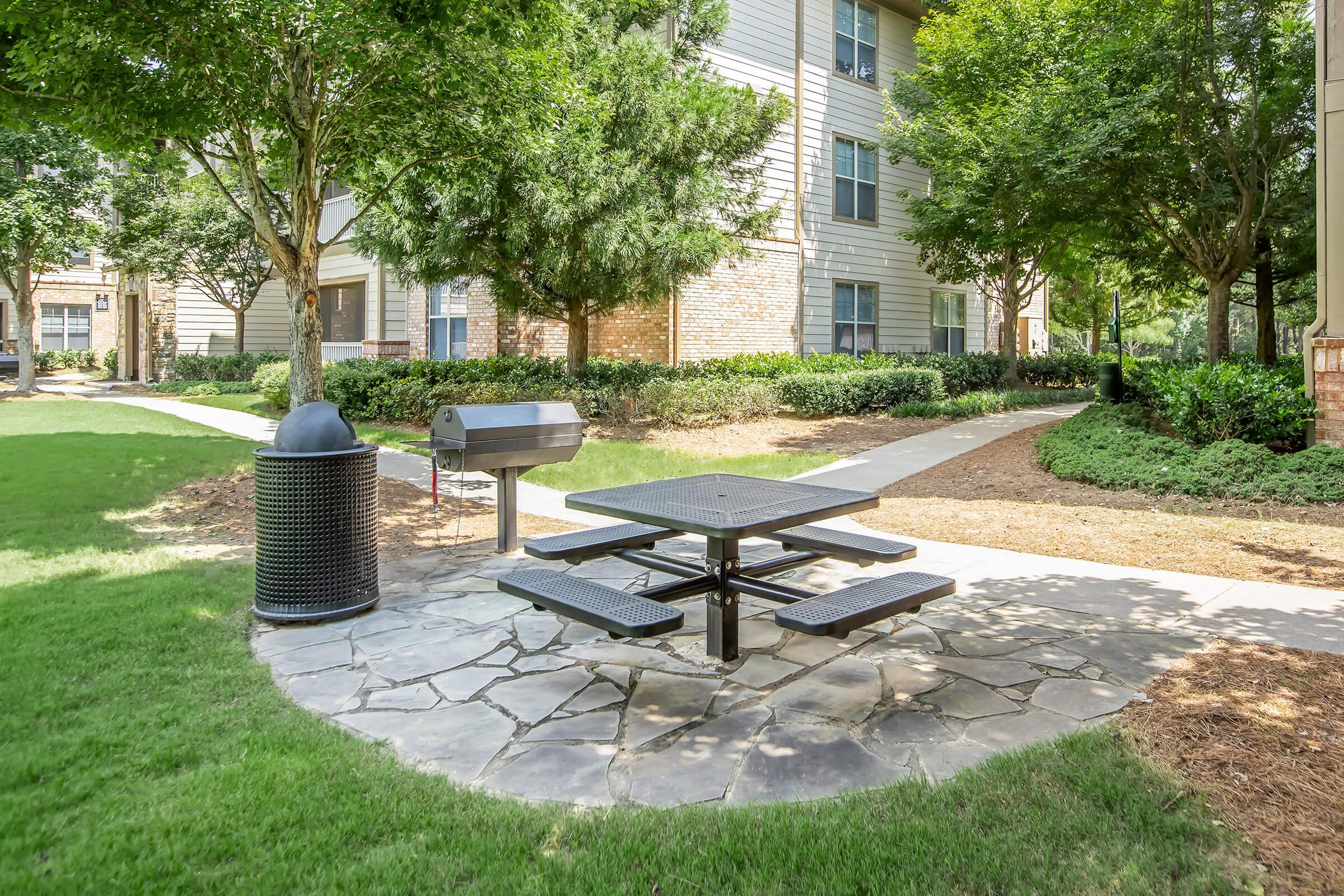 A shaded picnic area featuring a black metal table and benches, situated on a stone pathway in a landscaped courtyard. A charcoal grill is nearby, along with a black trash can. Surrounding the area are green lawns and trees, with apartment buildings visible in the background.