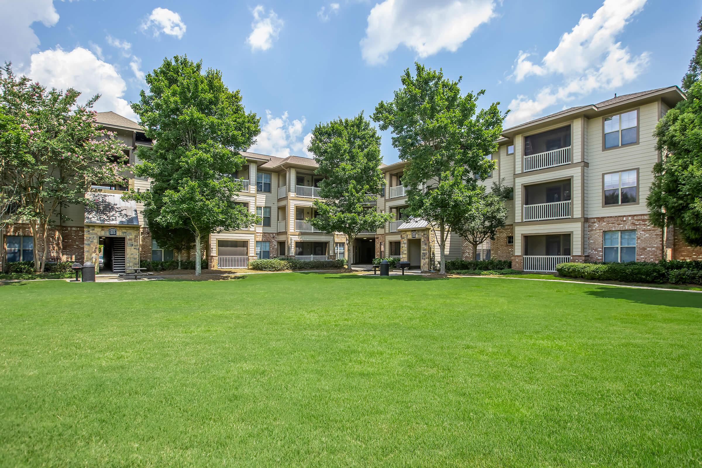 A well-maintained apartment complex with three-story buildings surrounded by lush green lawns and trees under a blue sky with fluffy clouds. The outdoor area features neatly trimmed grass and landscaping, creating a pleasant and inviting residential environment.