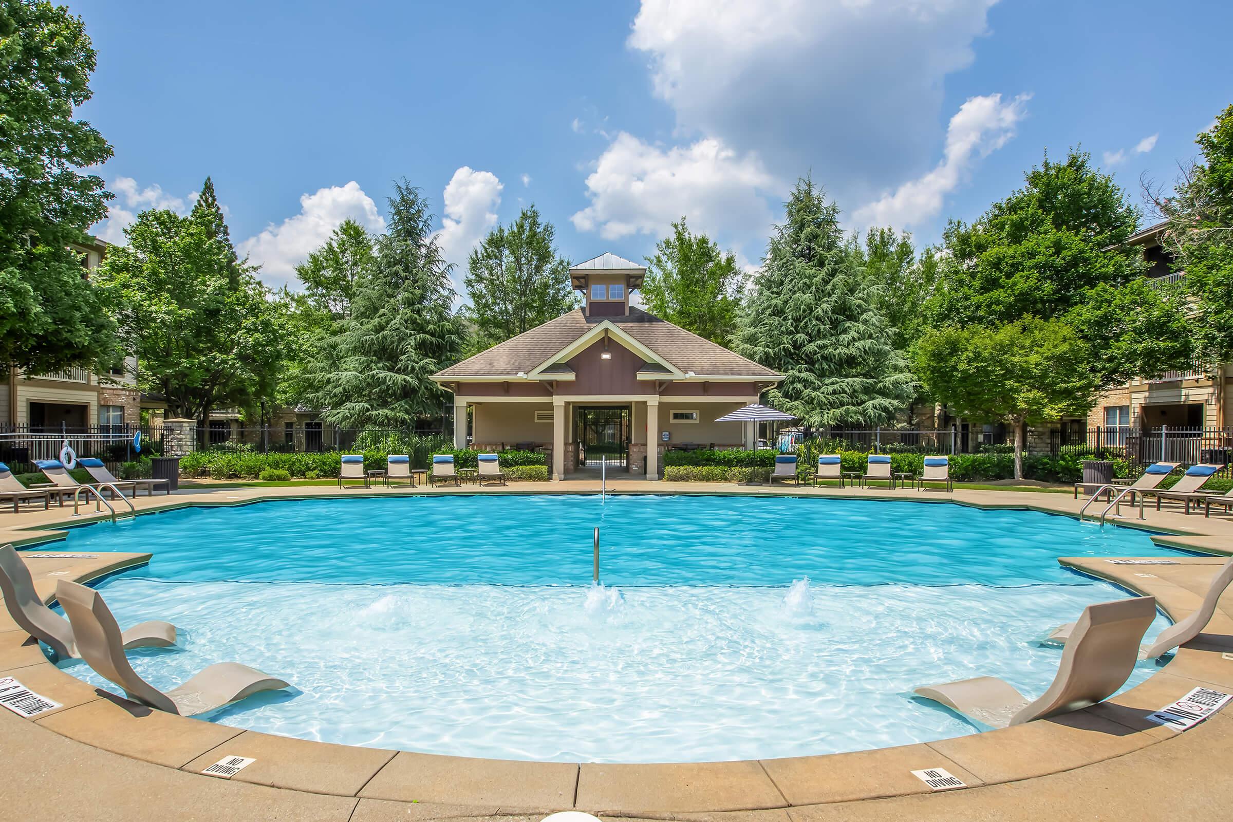 A spacious outdoor swimming pool with clear blue water, surrounded by lounge chairs. Lush green trees and well-maintained landscaping frame the area, with a building featuring a peaked roof in the background. The sky is bright and partly cloudy, creating a vibrant atmosphere.