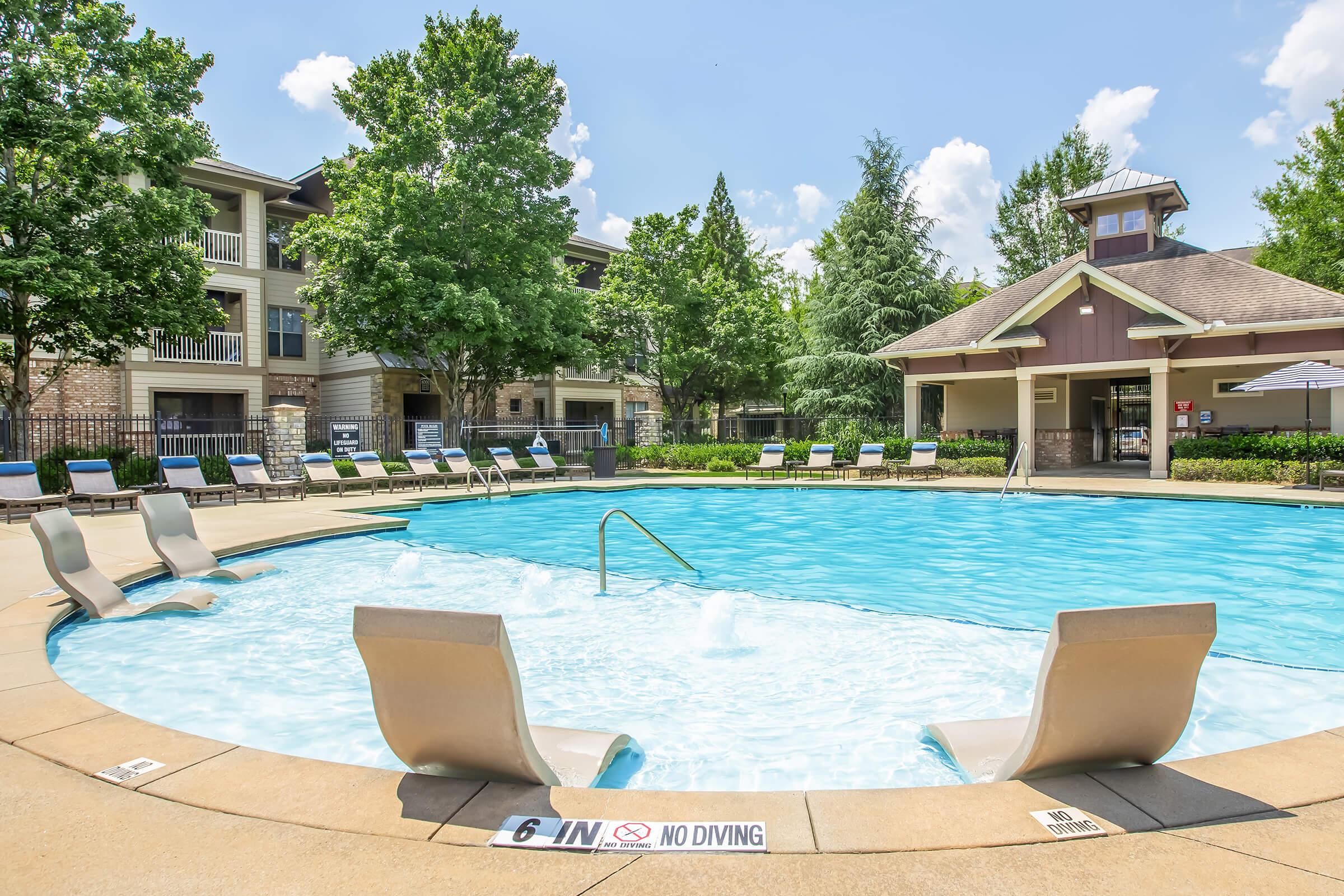A tranquil swimming pool surrounded by lounge chairs and greenery, with a shaded area in the background. The pool features a spa section and clear blue water, under a bright sky with fluffy clouds. Signs indicate "6 ft" depth and "No Diving" near the pool's edge.
