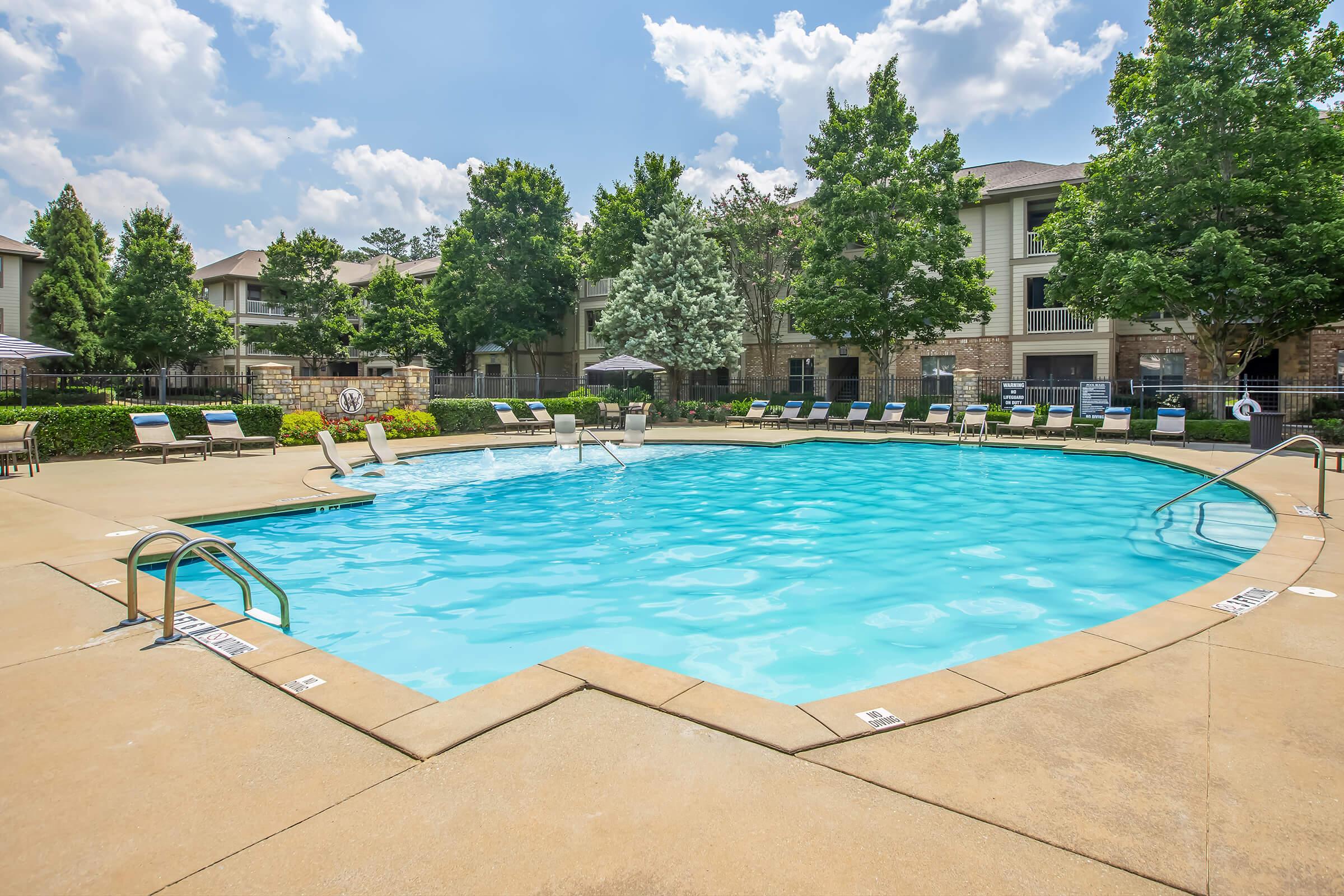 A clear blue swimming pool surrounded by lounge chairs and trees, set against an apartment complex. The sky is partly cloudy, creating a bright and inviting atmosphere. Nearby umbrellas provide shade, and the pool area is well-maintained, ideal for relaxation and recreation.
