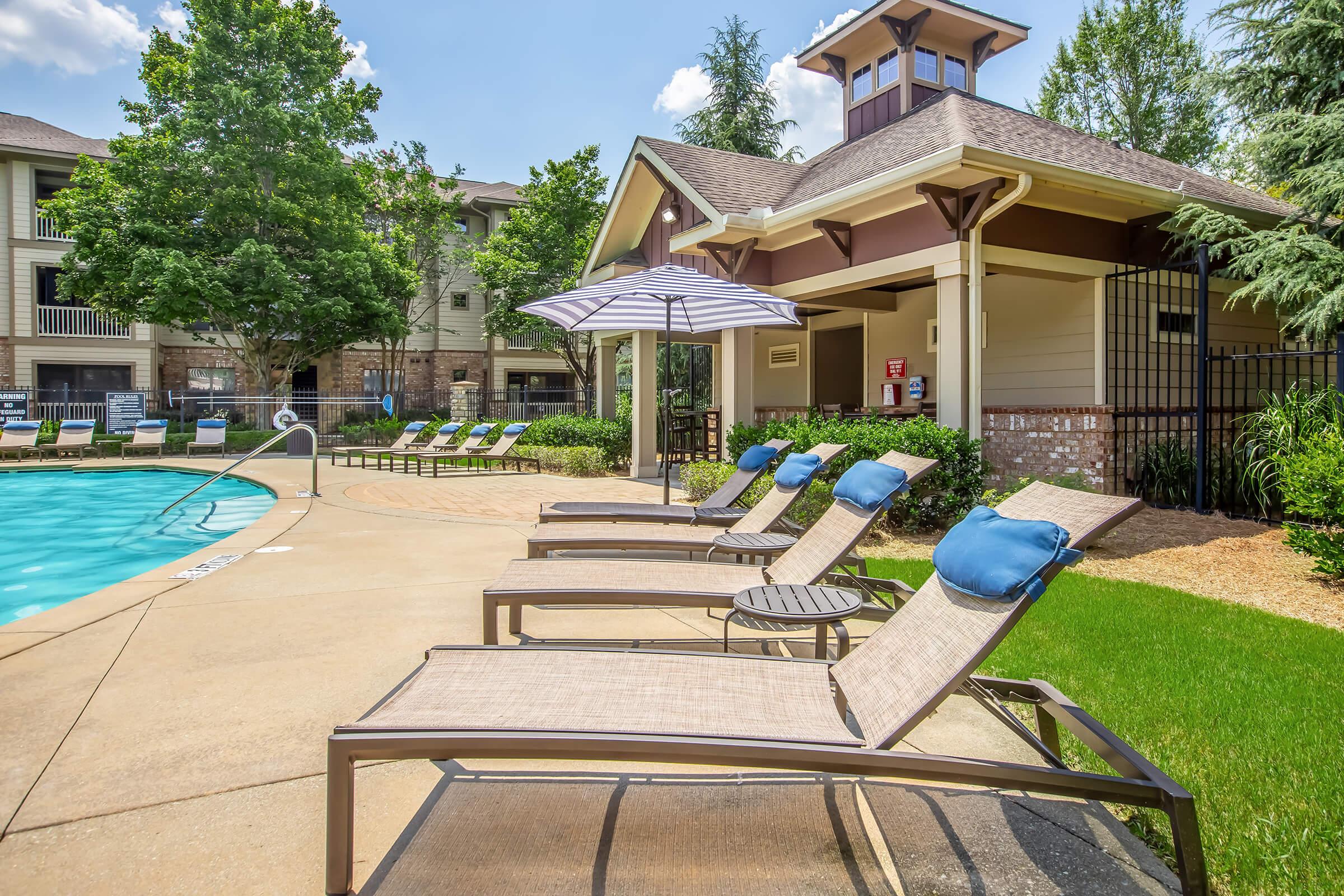 A tranquil outdoor scene featuring a swimming pool surrounded by lounge chairs and umbrellas. The pool area is bordered by lush green grass and trees, with an adjacent clubhouse structure. Bright blue skies complete the serene atmosphere.