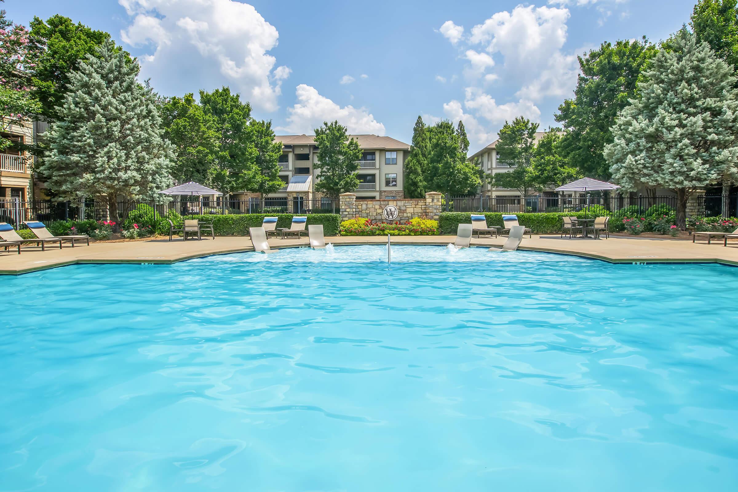 A sparkling blue pool surrounded by lounge chairs and sun umbrellas, with well-maintained greenery and blooming flowers around the perimeter. In the background, there are residential buildings under a clear blue sky with fluffy white clouds.