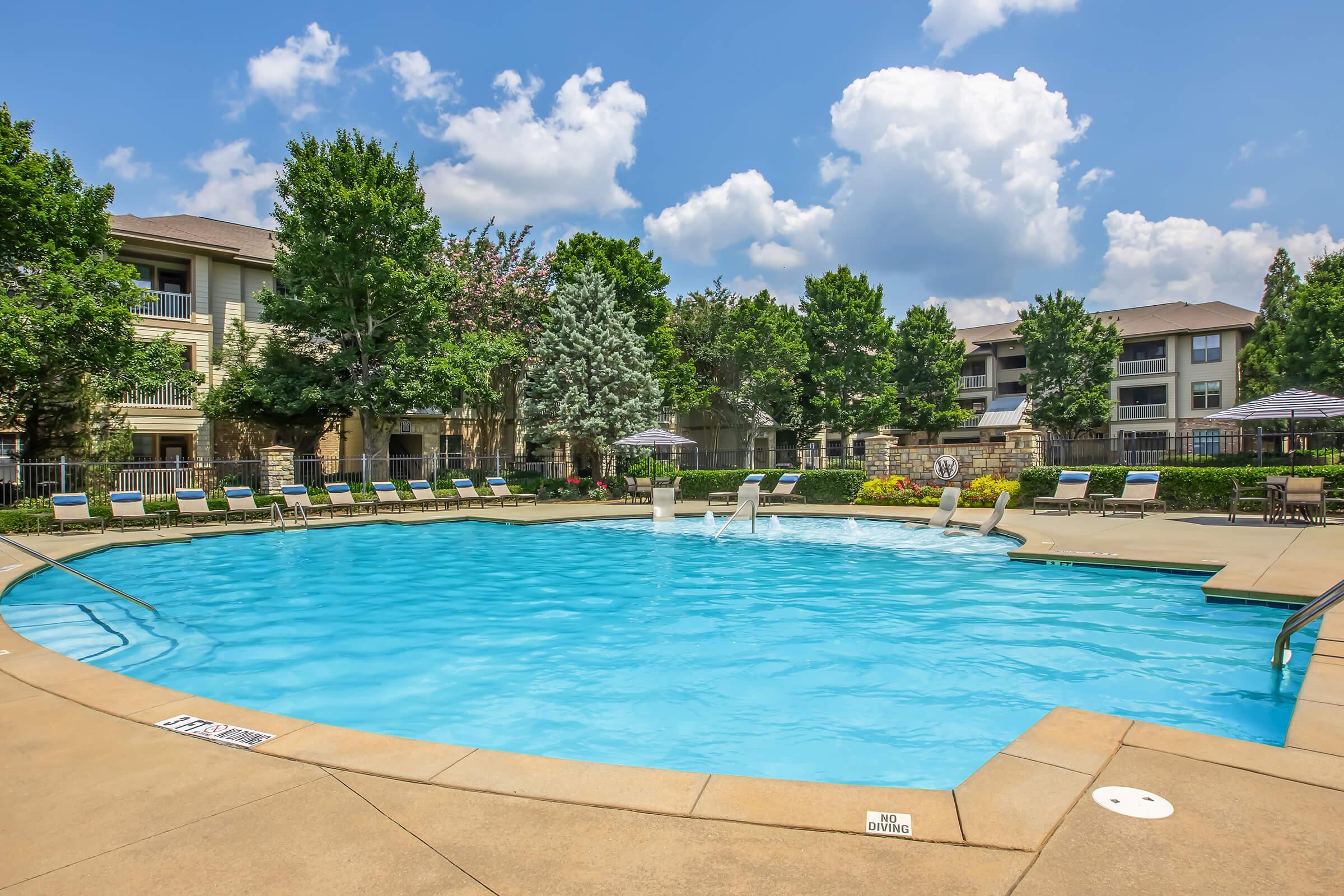 A sparkling swimming pool surrounded by lounge chairs and umbrellas, set in a landscaped area with trees and flowering plants. In the background, there are residential buildings under a blue sky with fluffy clouds, creating a relaxing outdoor atmosphere.