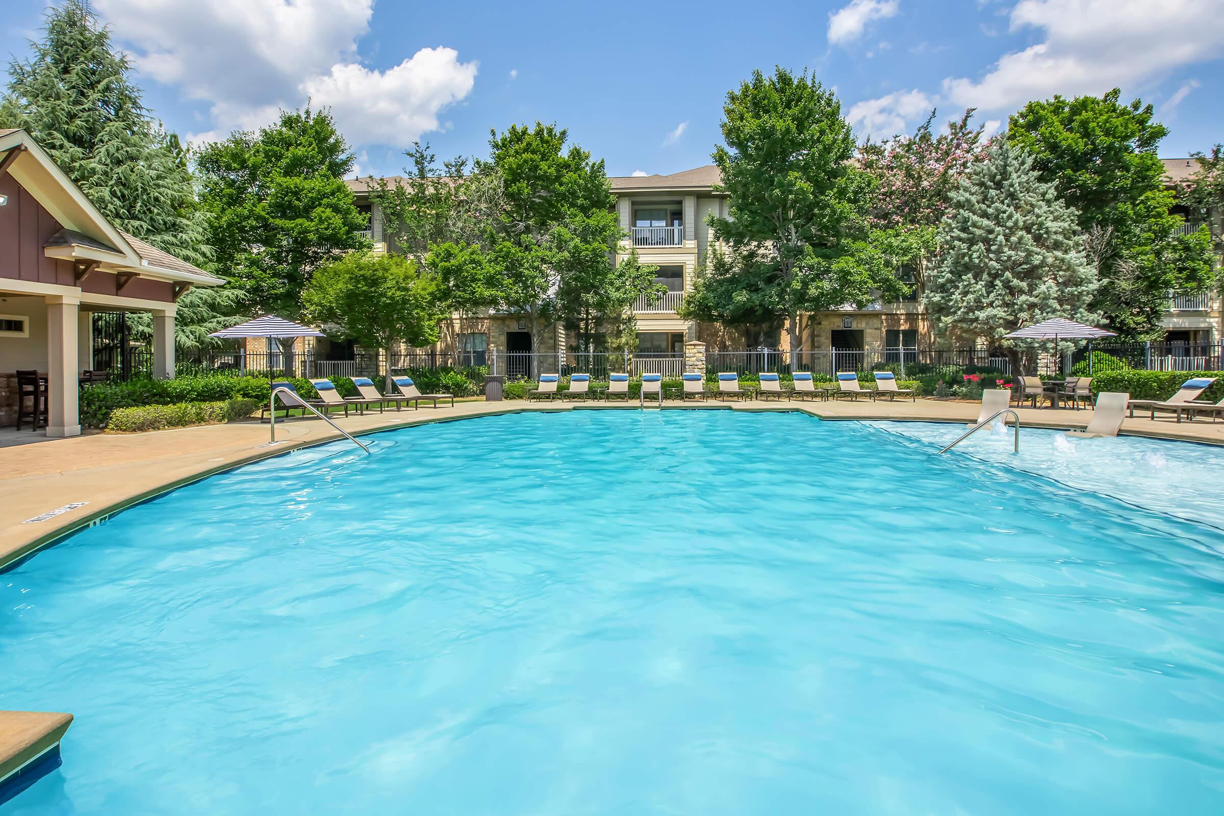 A clear blue swimming pool surrounded by lounge chairs, umbrellas, and lush greenery. Residential buildings are in the background, with sunny skies and a few clouds overhead, creating a relaxing outdoor atmosphere perfect for leisure.