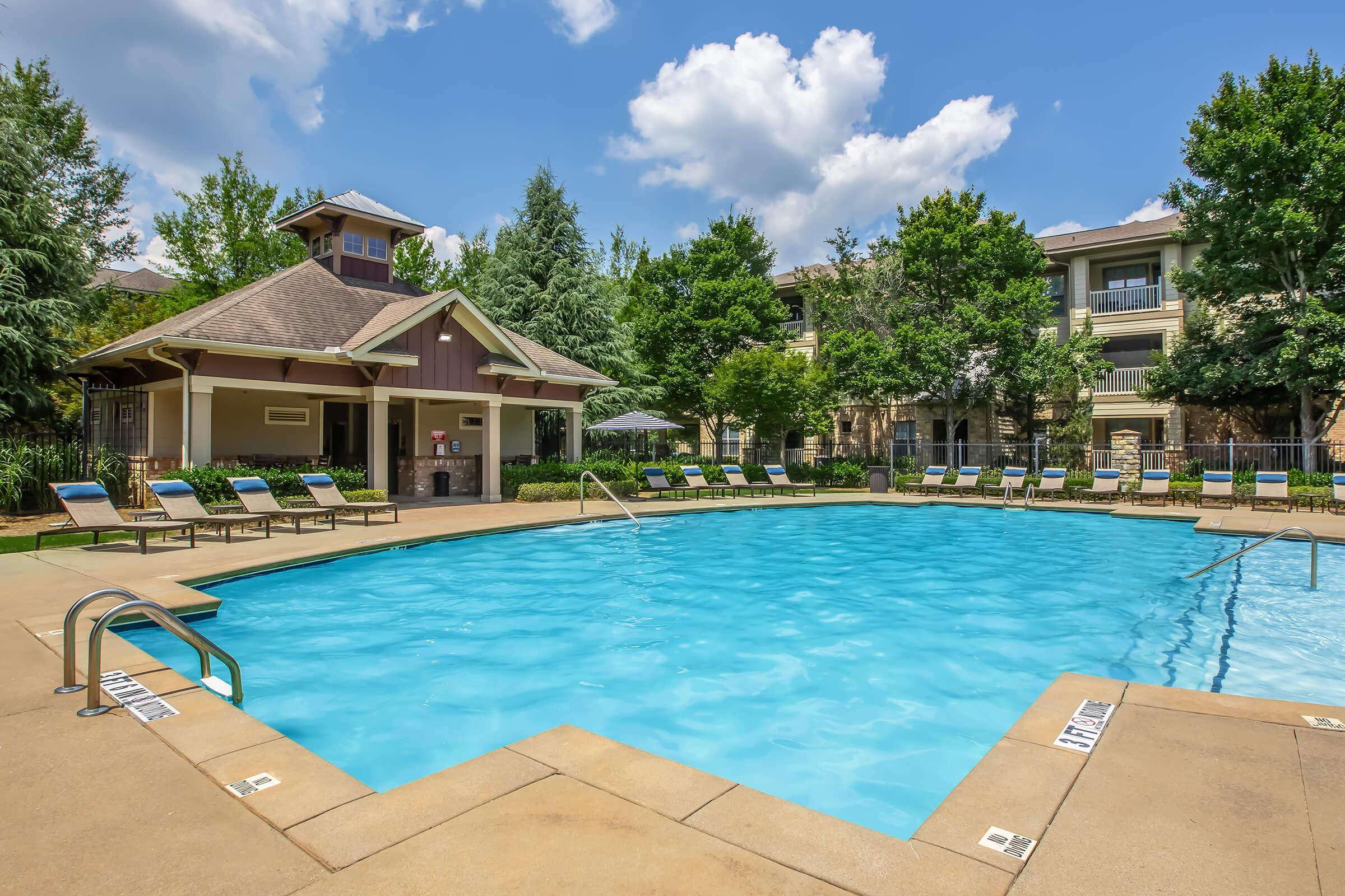 A clear blue swimming pool surrounded by lounge chairs and landscaped greenery. Nearby is a pool house with a gabled roof. The scene is set under a bright blue sky with fluffy white clouds, creating a relaxing and inviting atmosphere.