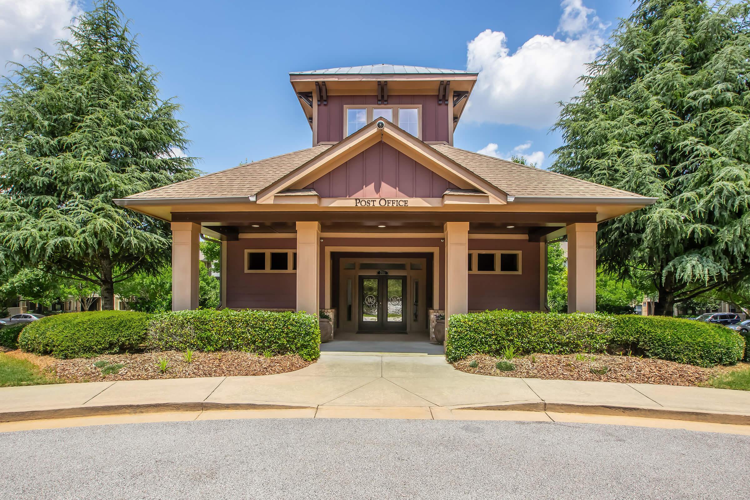 A modern, two-story building with a triangular roof, surrounded by manicured green shrubs and trees. The entrance features decorative columns and windows, with a circular driveway leading to the front. The sky is clear with a few clouds, indicating a bright, sunny day.