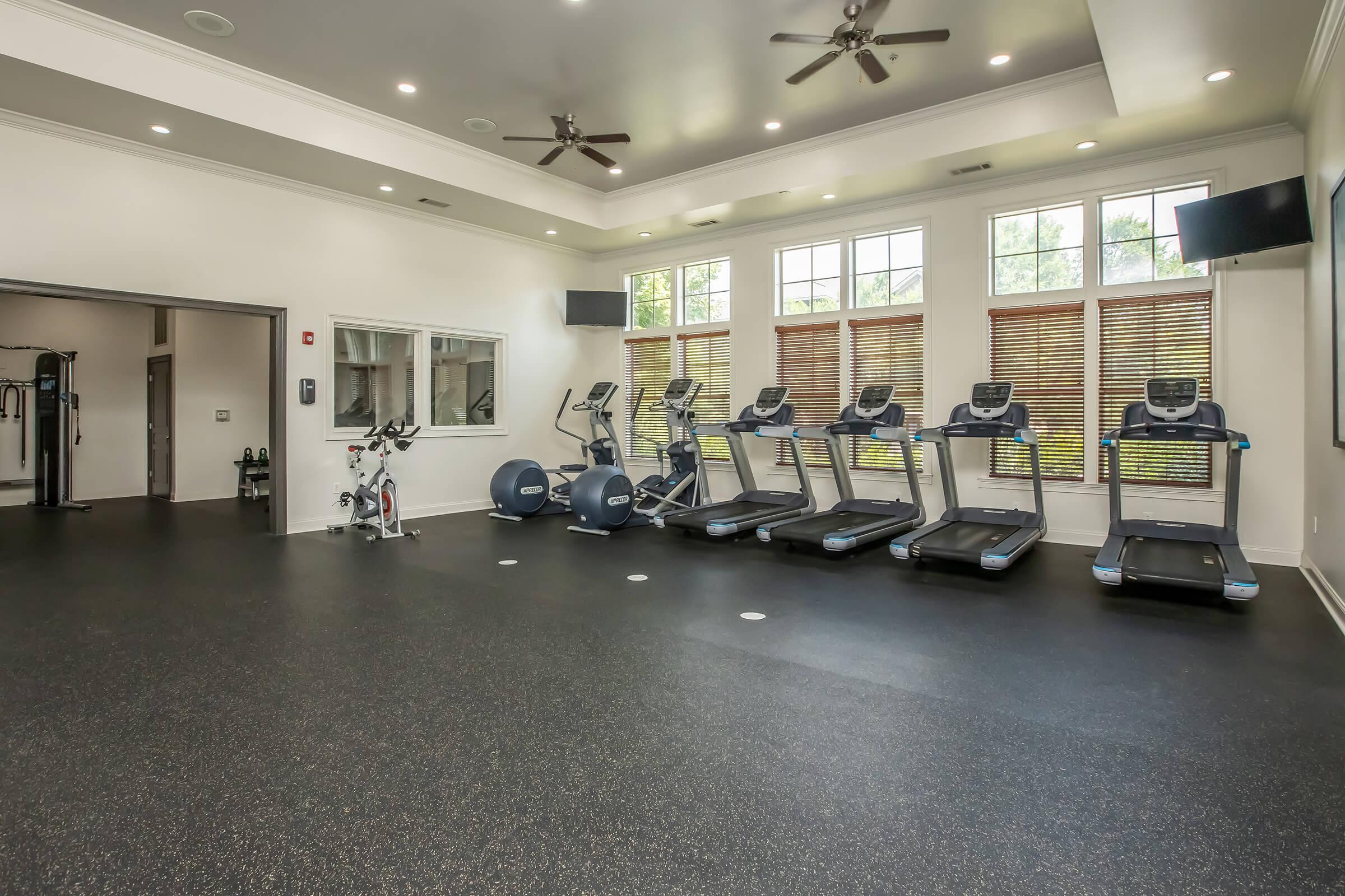 A modern fitness room featuring several exercise machines, including treadmills and a stationary bike. The space is well-lit with large windows and has a clean, polished floor. Fans are mounted on the ceiling, and there are screens on the walls, creating an inviting workout environment.