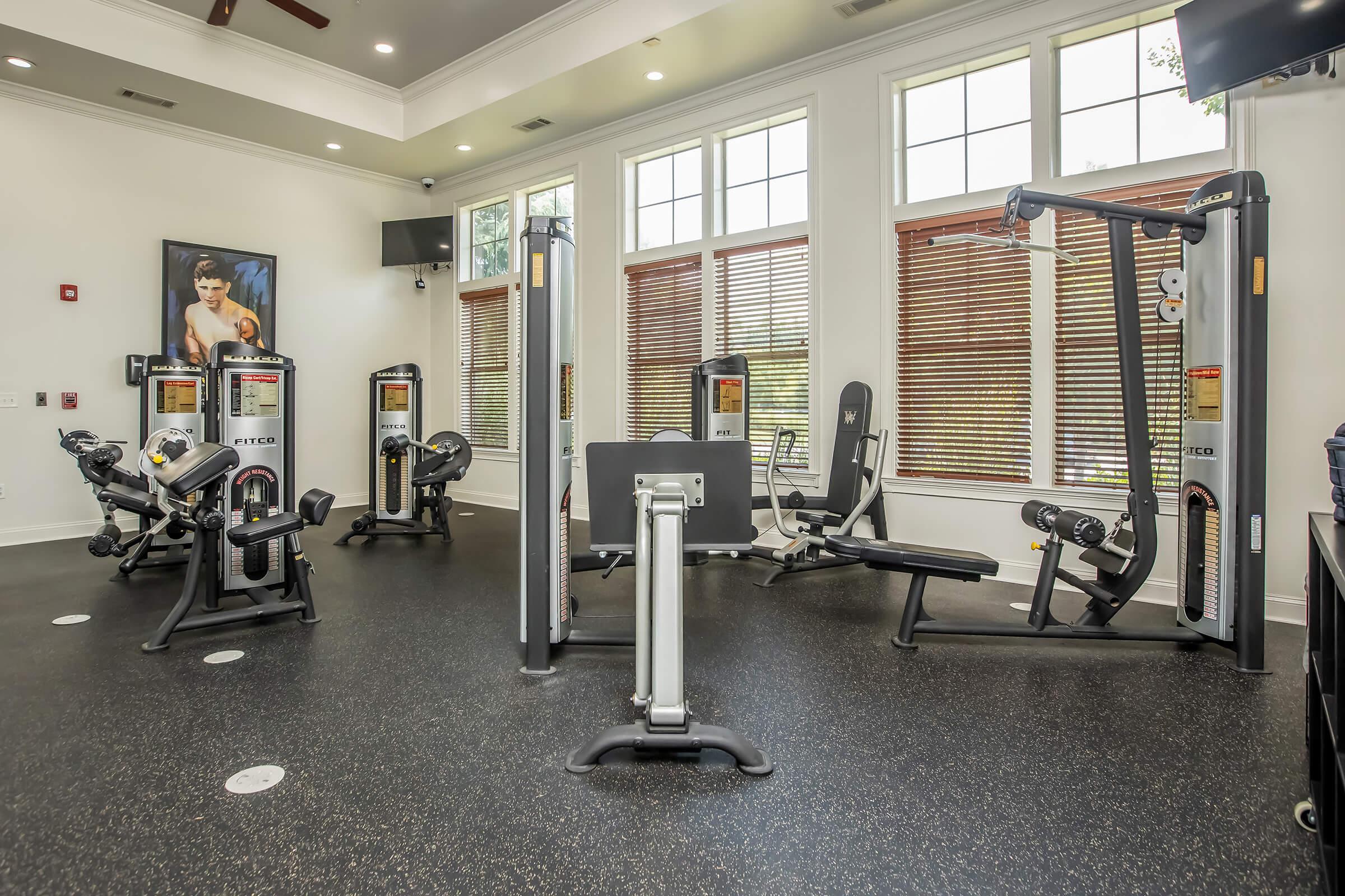 A well-lit gym interior featuring various exercise machines including weightlifting equipment and cardio machines. Large windows allow natural light, and there’s a motivational poster on the wall. The flooring is dark with a smooth finish, creating a modern workout space.