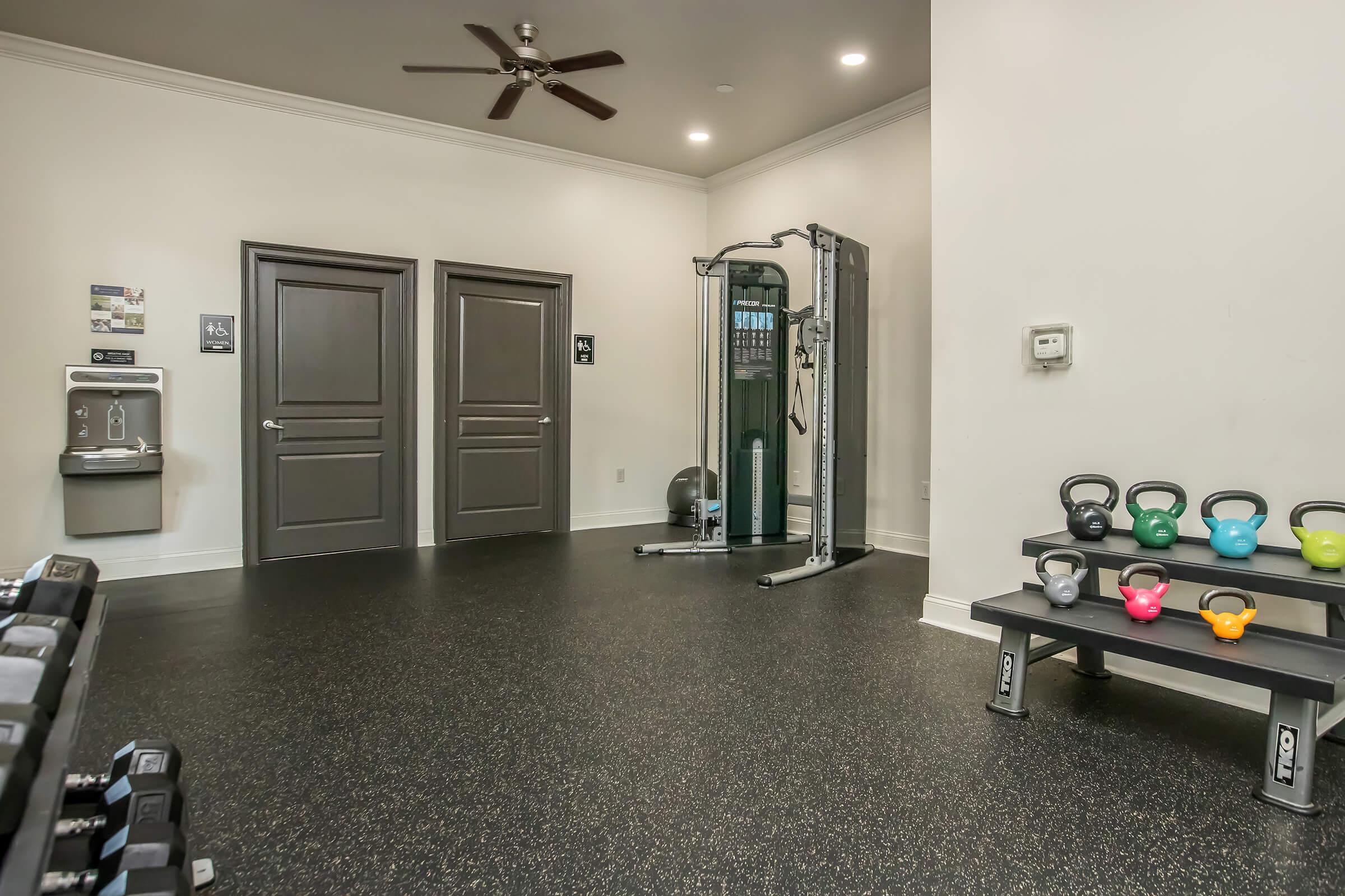 A fitness room featuring a range of weights, including dumbbells and kettlebells in various colors, a cable machine, and a water fountain. The walls are light-colored, and there are two closed doors visible. The flooring is dark and rubberized, providing a comfortable workout environment.