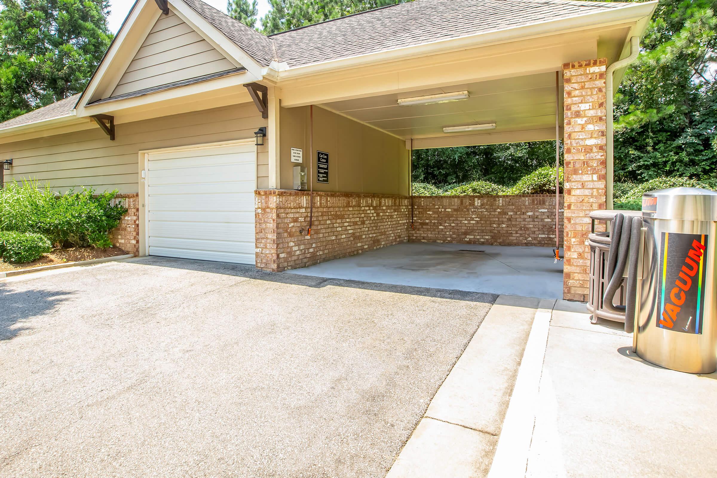 A parking garage with a brick wall and a clean concrete floor. There is a vacuum station on the right side and a garage door on the front. Lush greenery is visible in the background, providing a serene atmosphere. The area is well-lit and organized.