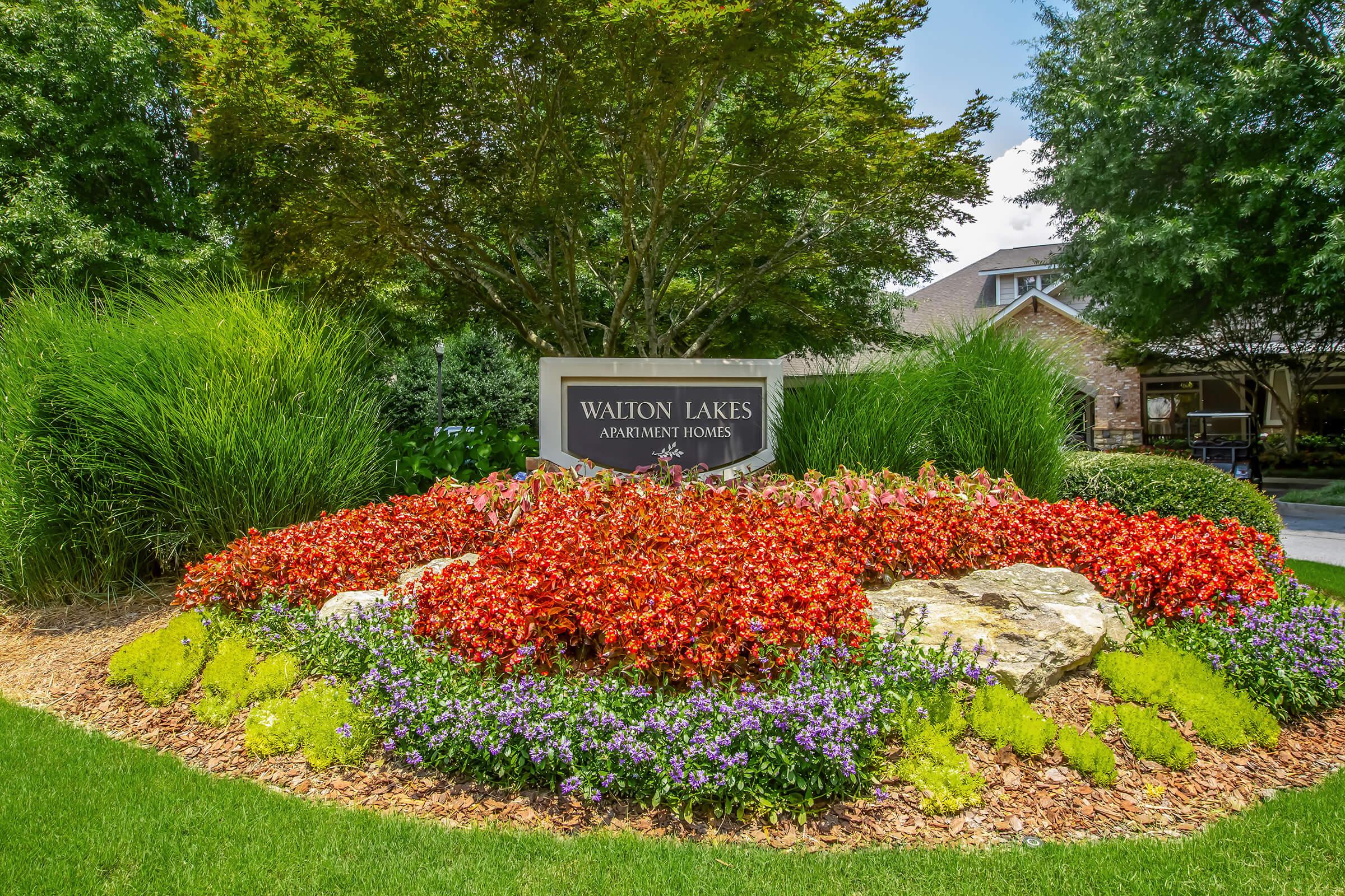 A vibrant flower bed filled with red, purple, and yellow flowers surrounds a sign that reads "Walton Lakes Apartment Homes." Lush green grass and trees are visible in the background, creating a welcoming outdoor atmosphere.