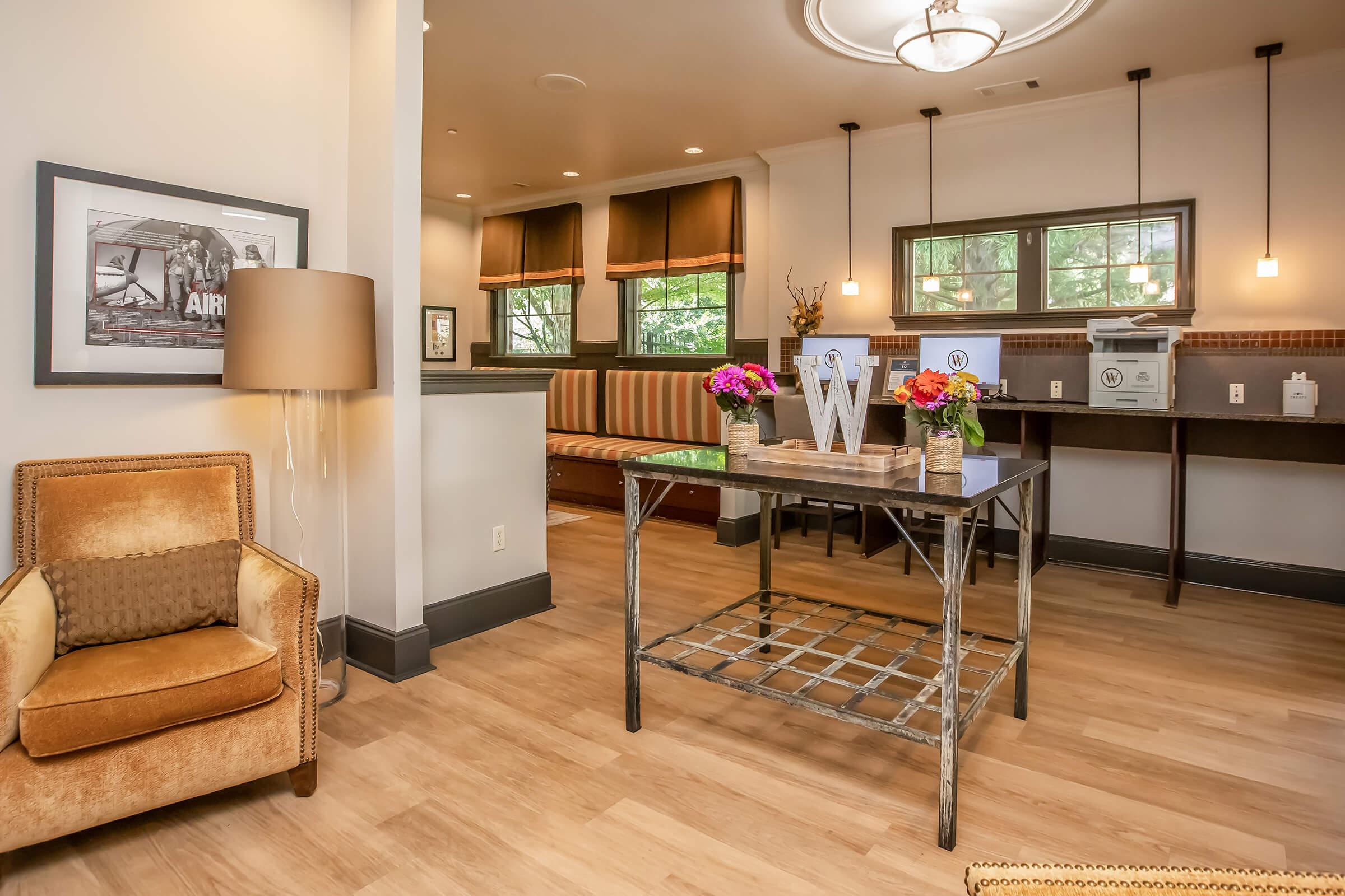 Cozy interior of a sitting area featuring a comfortable orange armchair, a metal table with decorative items, and a wall-mounted display. Large windows with brown curtains allow natural light, and there is a framed picture on the wall. The space has wooden flooring and a warm, inviting atmosphere.