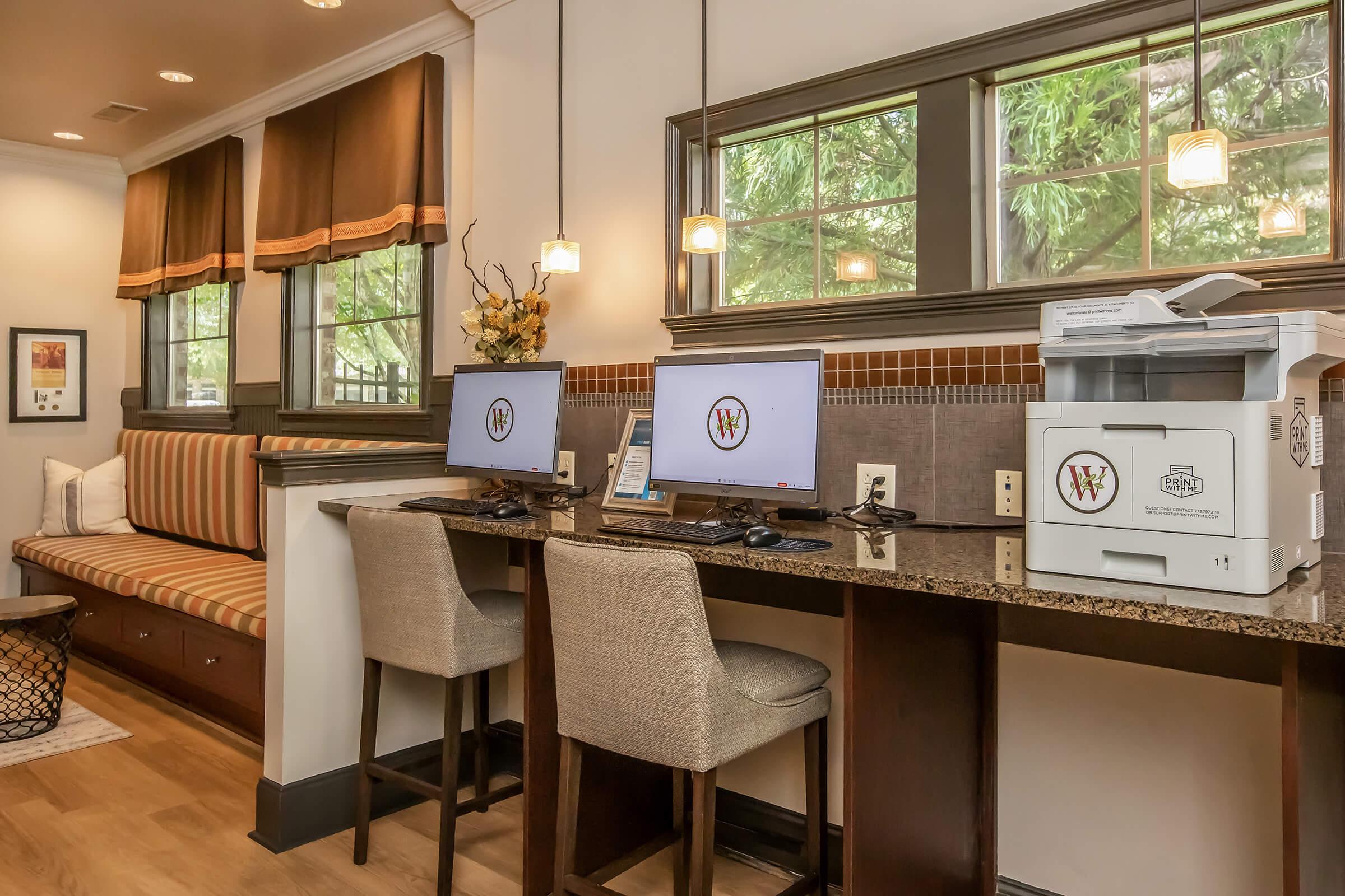 A cozy workspace featuring two computers on a granite counter, with a printer nearby. There is a comfortable seating area with striped cushions, large windows allowing natural light, and decorative elements like plants and artwork. The space is designed for productivity and comfort.